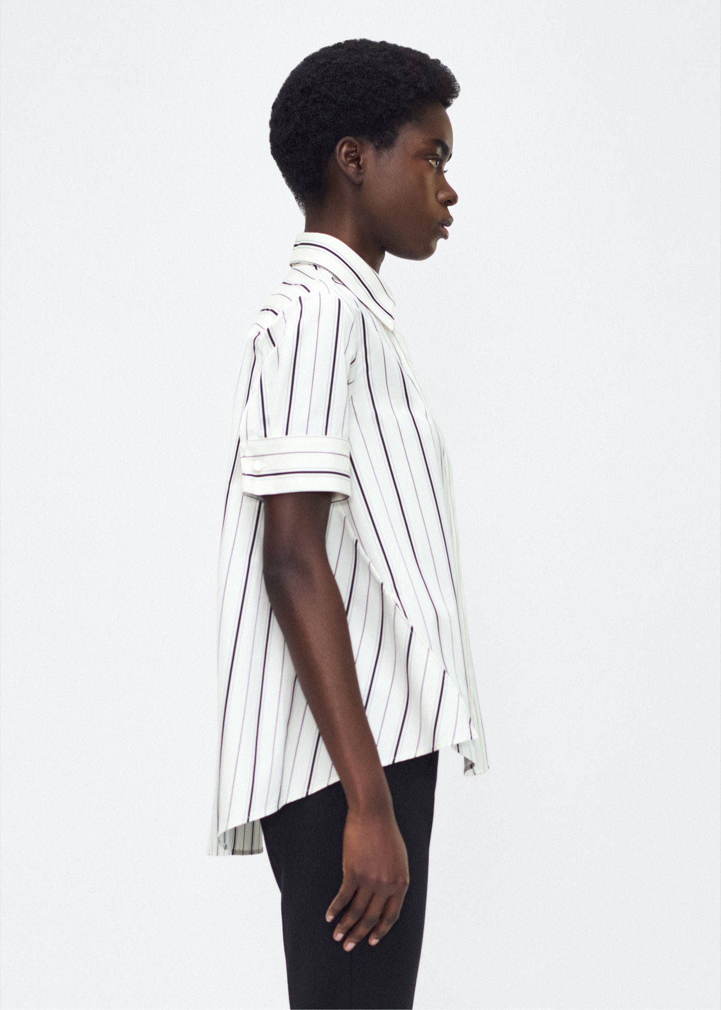 Model side facing image – Trapeze Shirt, P25106SS, Ivory/Black – Adam Lippes, P25 collection: The Adam Lippes Trapeze Shirt remains a timeless favorite from the atelier. Crafted from crisp Italian cotton, this season’s version features a classic ivory and black stripe. The piece features diamond monogram mother of pearl buttons along the center front placket, collar stand and French cuffs. Designed with side slits for a breezy, relaxed fit.