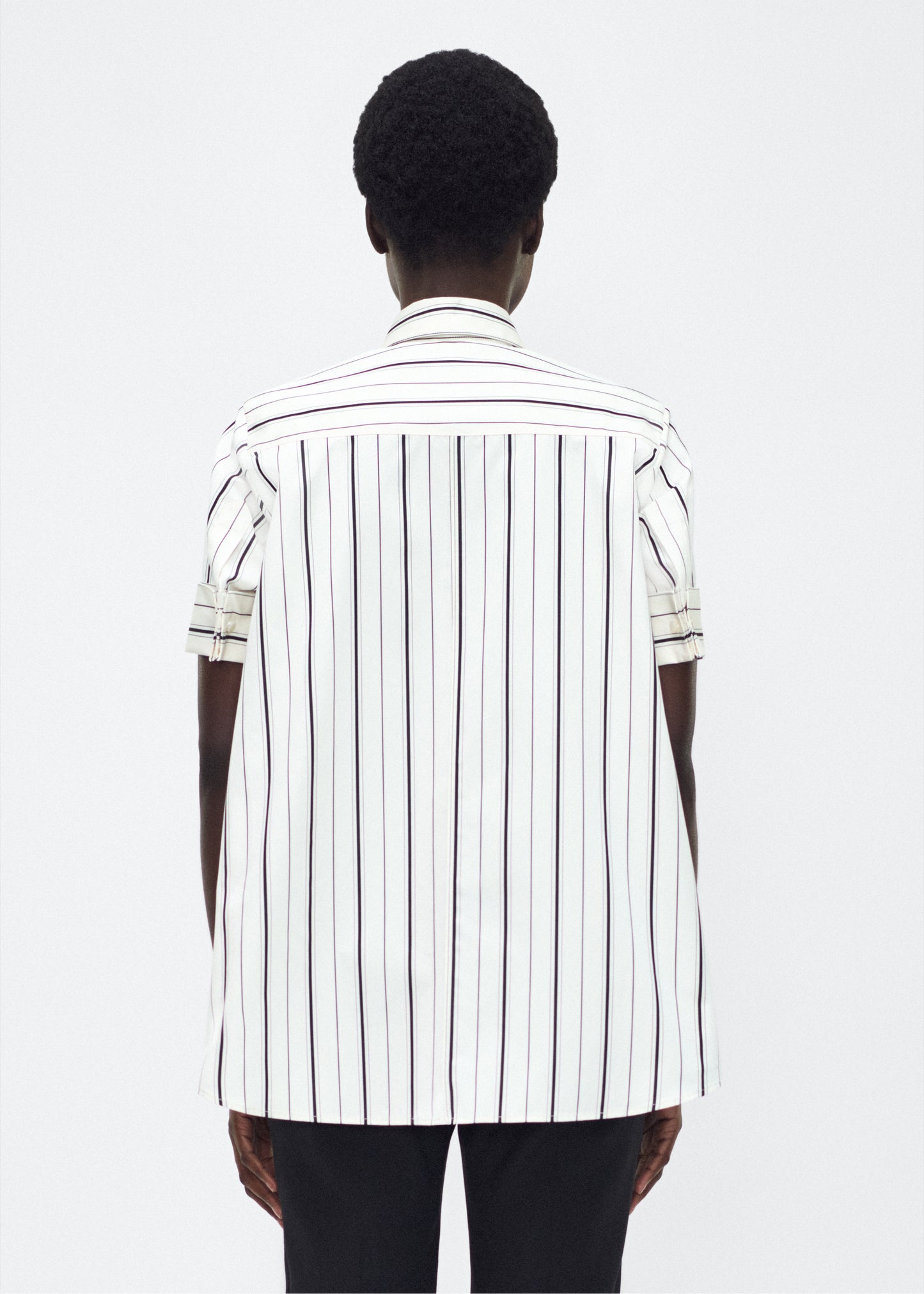Model back facing image – Trapeze Shirt, P25106SS, Ivory/Black – Adam Lippes, P25 collection: The Adam Lippes Trapeze Shirt remains a timeless favorite from the atelier. Crafted from crisp Italian cotton, this season’s version features a classic ivory and black stripe. The piece features diamond monogram mother of pearl buttons along the center front placket, collar stand and French cuffs. Designed with side slits for a breezy, relaxed fit.