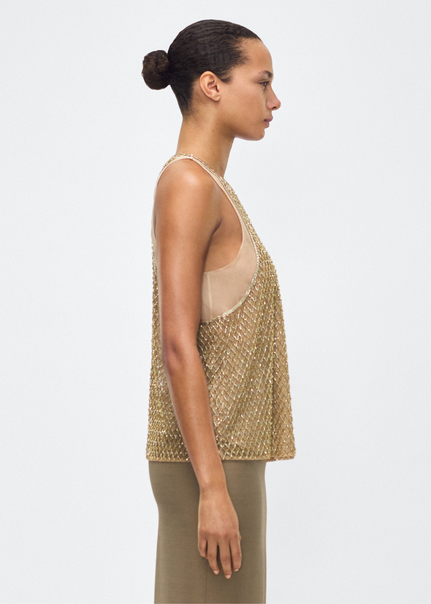Image of model facing side – Fable Top, P25110TU, Gold – Adam Lippes, P25 collection: The Fable Top in beaded tulle features intricate glass beading, a meticulous hand-done process that takes several days, arranged in the signature diamond motif for a striking visual effect. This new shape from the atelier combines fine craftsmanship with a clean, modern silhouette.