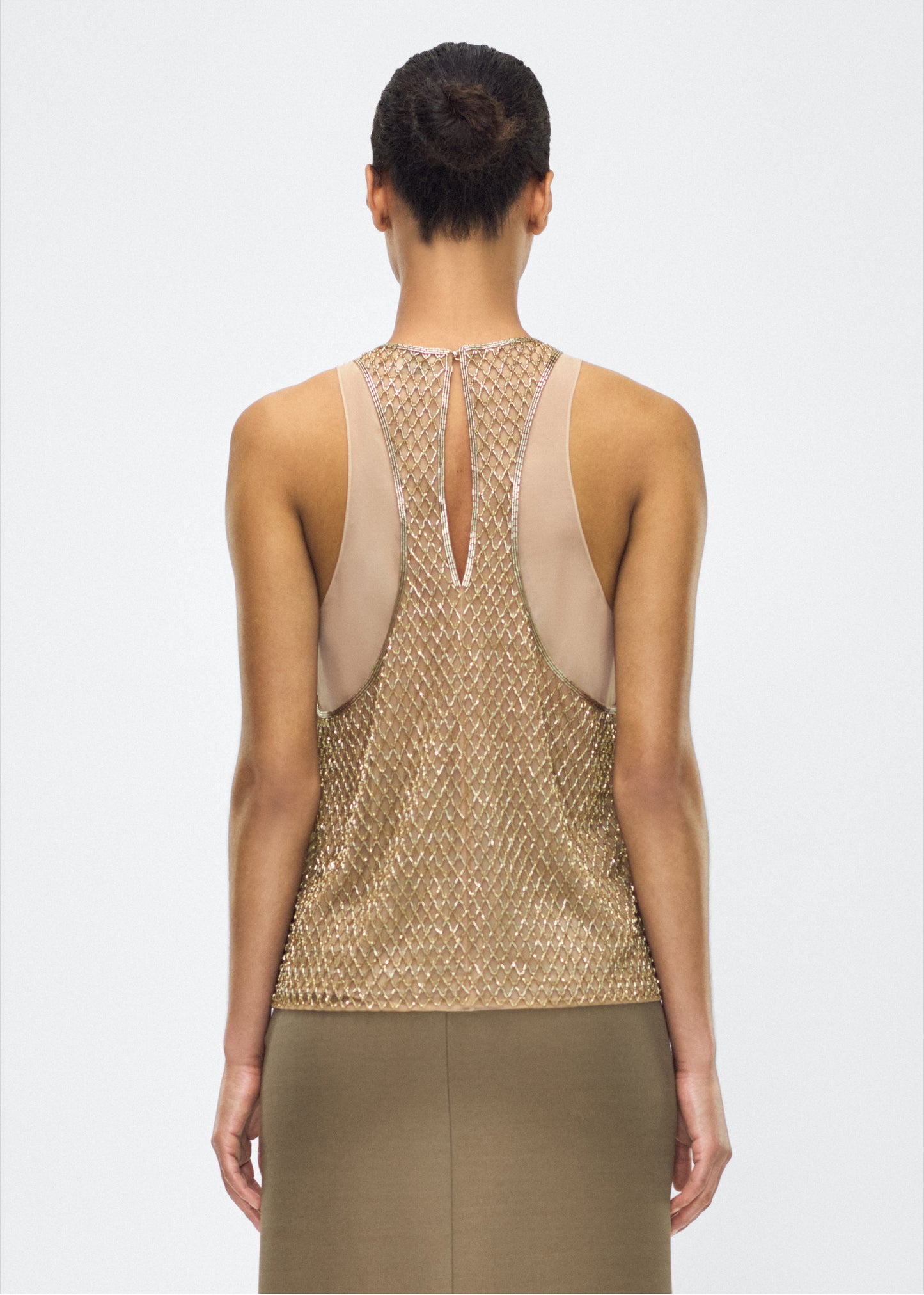 Image of model facing back – Fable Top, P25110TU, Gold – Adam Lippes, P25 collection: The Fable Top in beaded tulle features intricate glass beading, a meticulous hand-done process that takes several days, arranged in the signature diamond motif for a striking visual effect. This new shape from the atelier combines fine craftsmanship with a clean, modern silhouette.