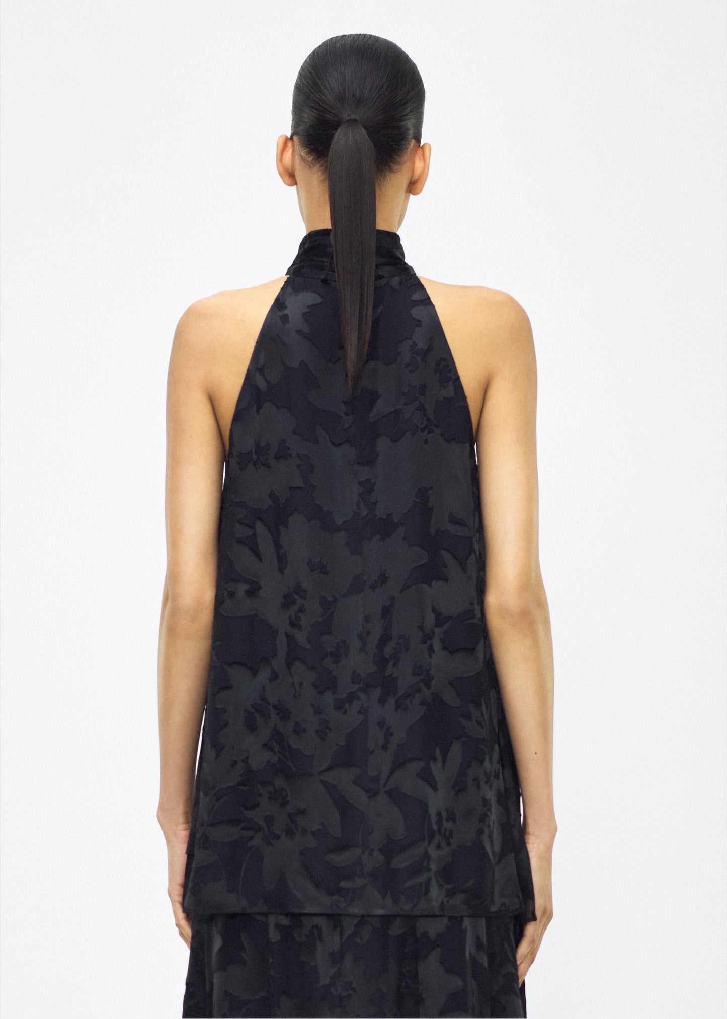 Image of model facing back – giulia top, P25117DV, Black – Adam Lippes, P25 collection: The Giulia Top is crafted from elegant burn-out fabric, and features an intricate floral design. The classic black hue enhances the rich texture, while the flowing silhouette exudes sophistication. Pair with the Emi Skirt in Devore for a coordinated ensemble.