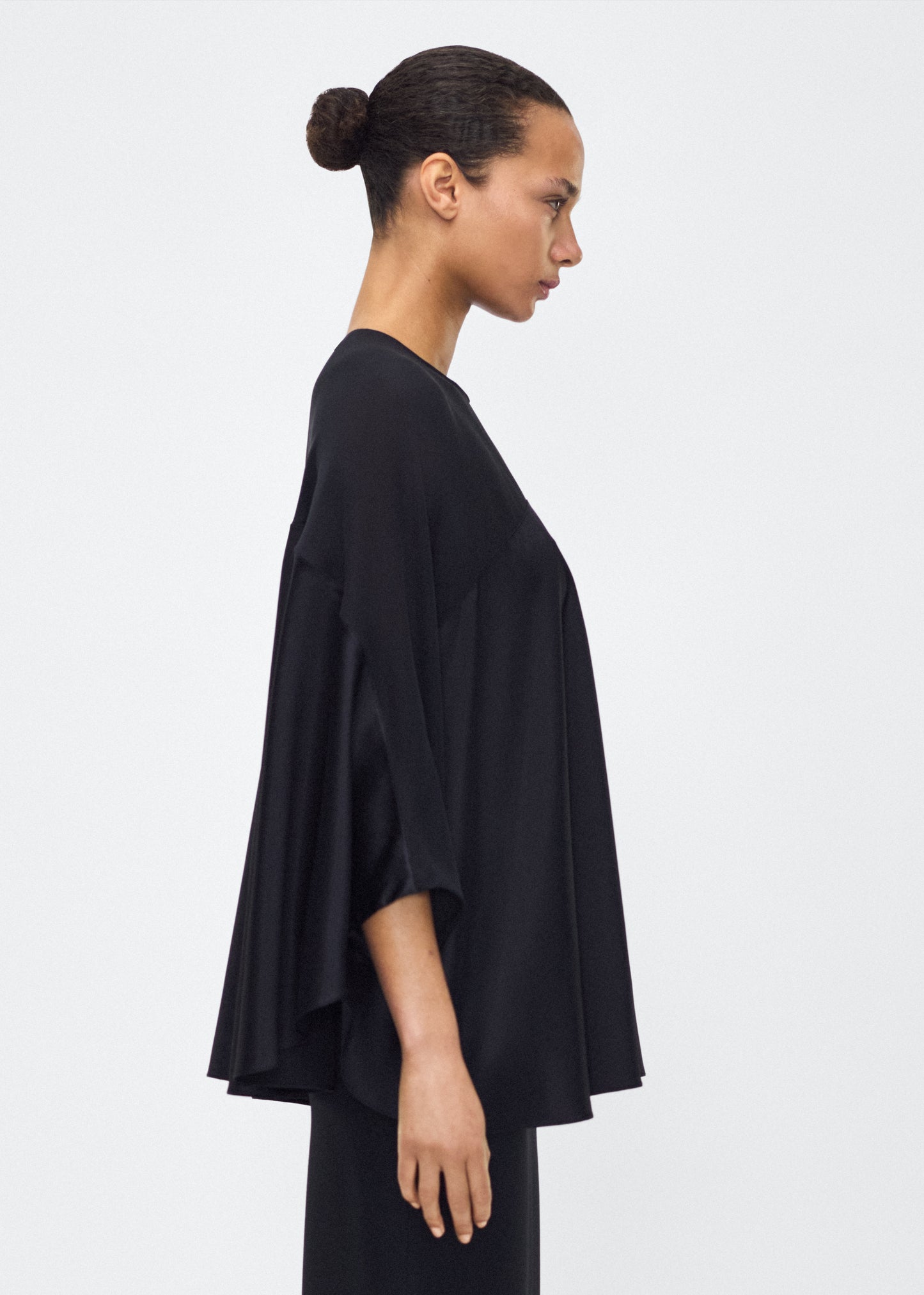 Image of model facing side wearing the apollo top, P25127DC, black – Adam Lippes, P25 collection: A new shape from the atelier, the Apollo Top is crafted from luxurious 100% silk in a timeless black hue. The flowing, draped design features a high-neck collar and a cascading silhouette, exuding modern elegance.