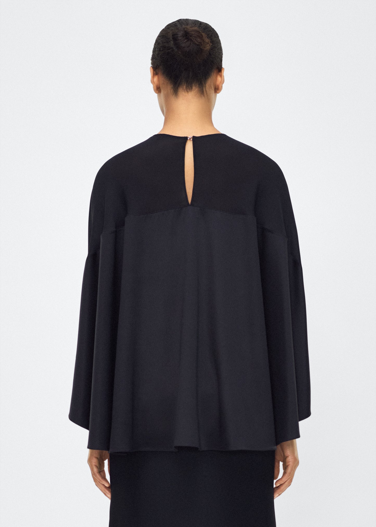 Image of model facing back wearing the apollo top, P25127DC, black – Adam Lippes, P25 collection: A new shape from the atelier, the Apollo Top is crafted from luxurious 100% silk in a timeless black hue. The flowing, draped design features a high-neck collar and a cascading silhouette, exuding modern elegance.