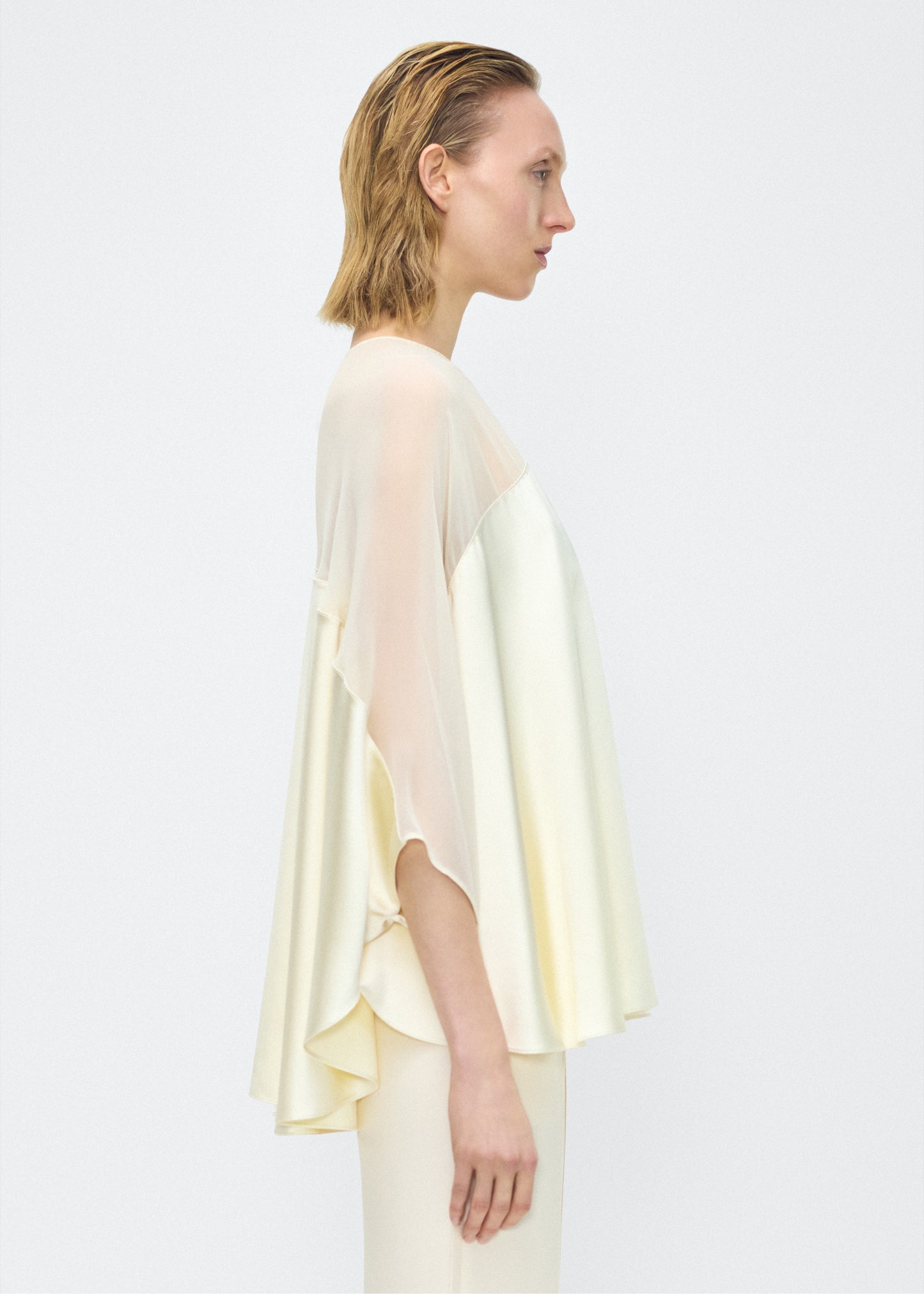 Model facing side – apollo top, P25127DC, cream – Adam Lippes, P25 collection: A new shape from the atelier, the Apollo Top is crafted from luxurious 100% silk in a soft cream hue. The flowing, draped design features a high-neck collar and a cascading silhouette, exuding modern elegance.