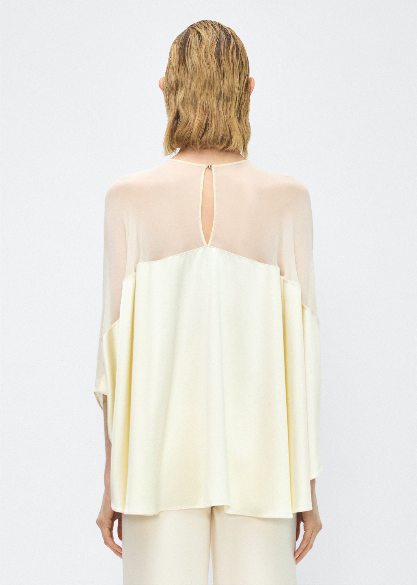 Model facing back – apollo top, P25127DC, cream – Adam Lippes, P25 collection: A new shape from the atelier, the Apollo Top is crafted from luxurious 100% silk in a soft cream hue. The flowing, draped design features a high-neck collar and a cascading silhouette, exuding modern elegance.