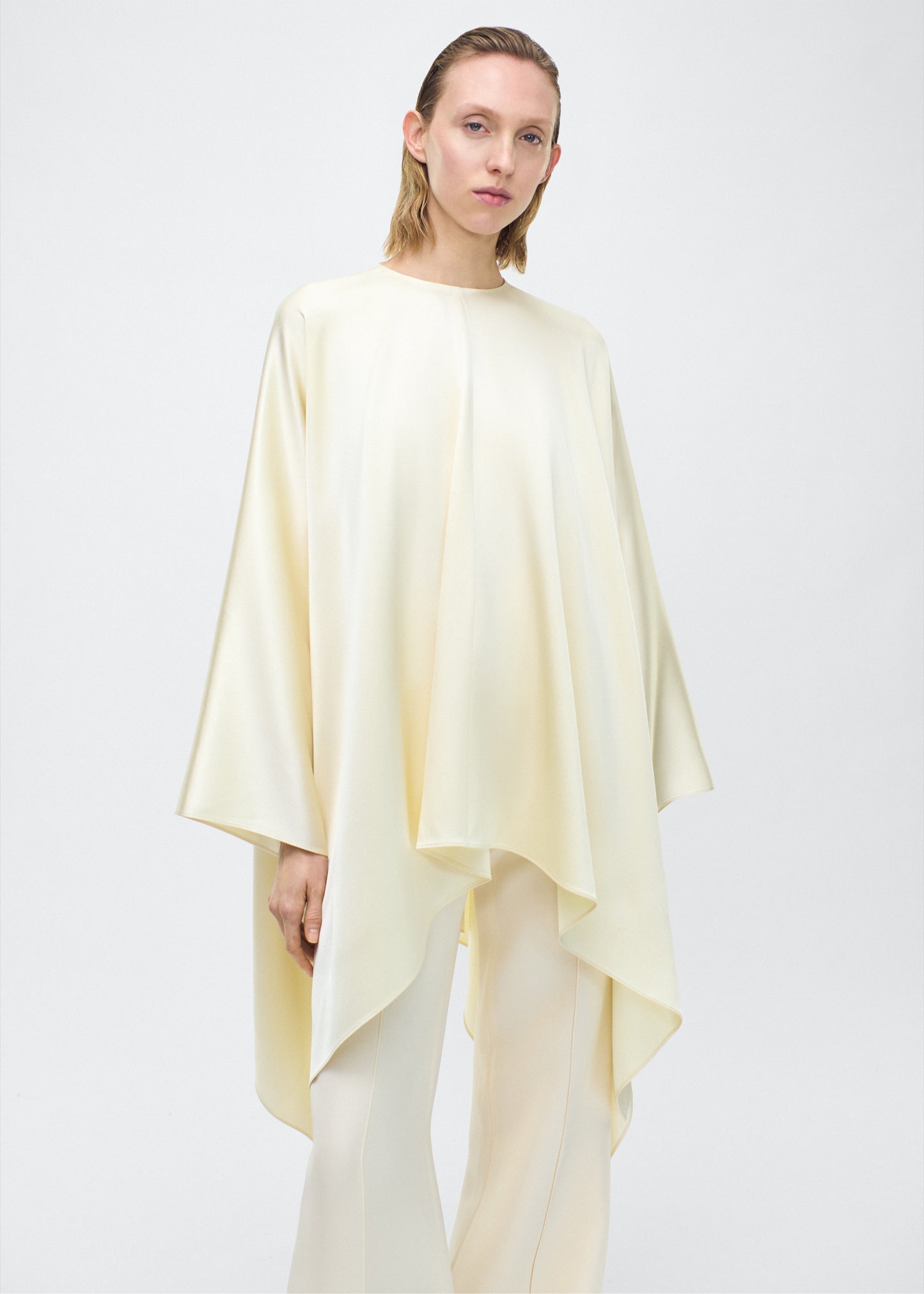 Model facing front angled – tana top , P25134HM, cream – Adam Lippes, P25 collection: The Tana Top is crafted from luxurious double-face charmeuse, offering a rich, fluid drape in a delicate cream hue. Featuring a detachable scarf for versatile styling options. The clean lines and elegant shape make it a statement piece, perfect for both dressing up and down.