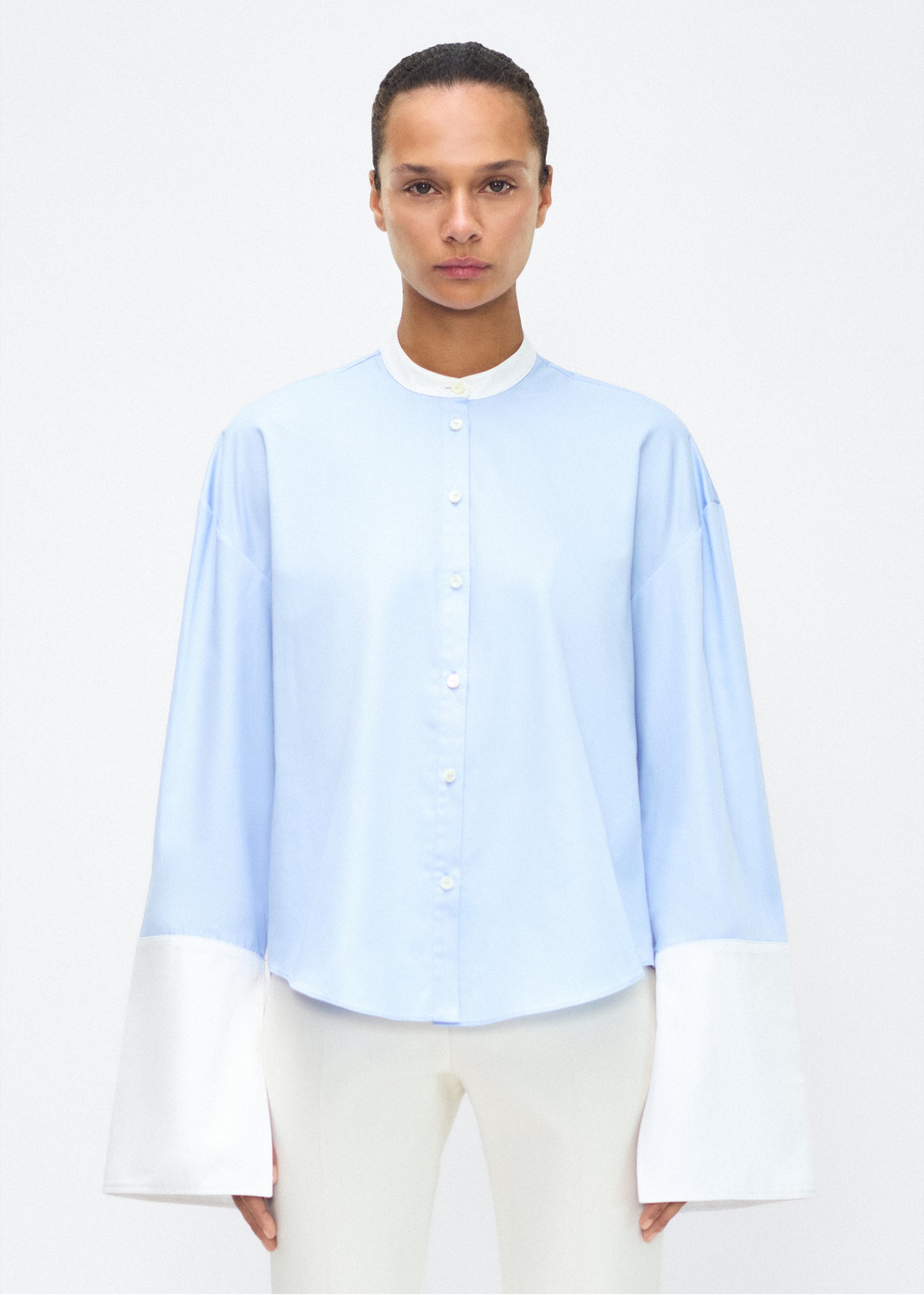 Model facing front – bram top, P25139CS, Pale Blue – Adam Lippes, P25 collection: A new shape from the atelier, the Bram Top is crafted from soft organic cotton poplin in a refreshing pale blue hue. The relaxed fit i enhanced by contrasting white cuffs and collar, with a subtle slit near the cuffs for added detail. Made from the highest quality cotton and featuring diamond monogram mother of pearl buttons down the center front, the top offers elegance and comfort.