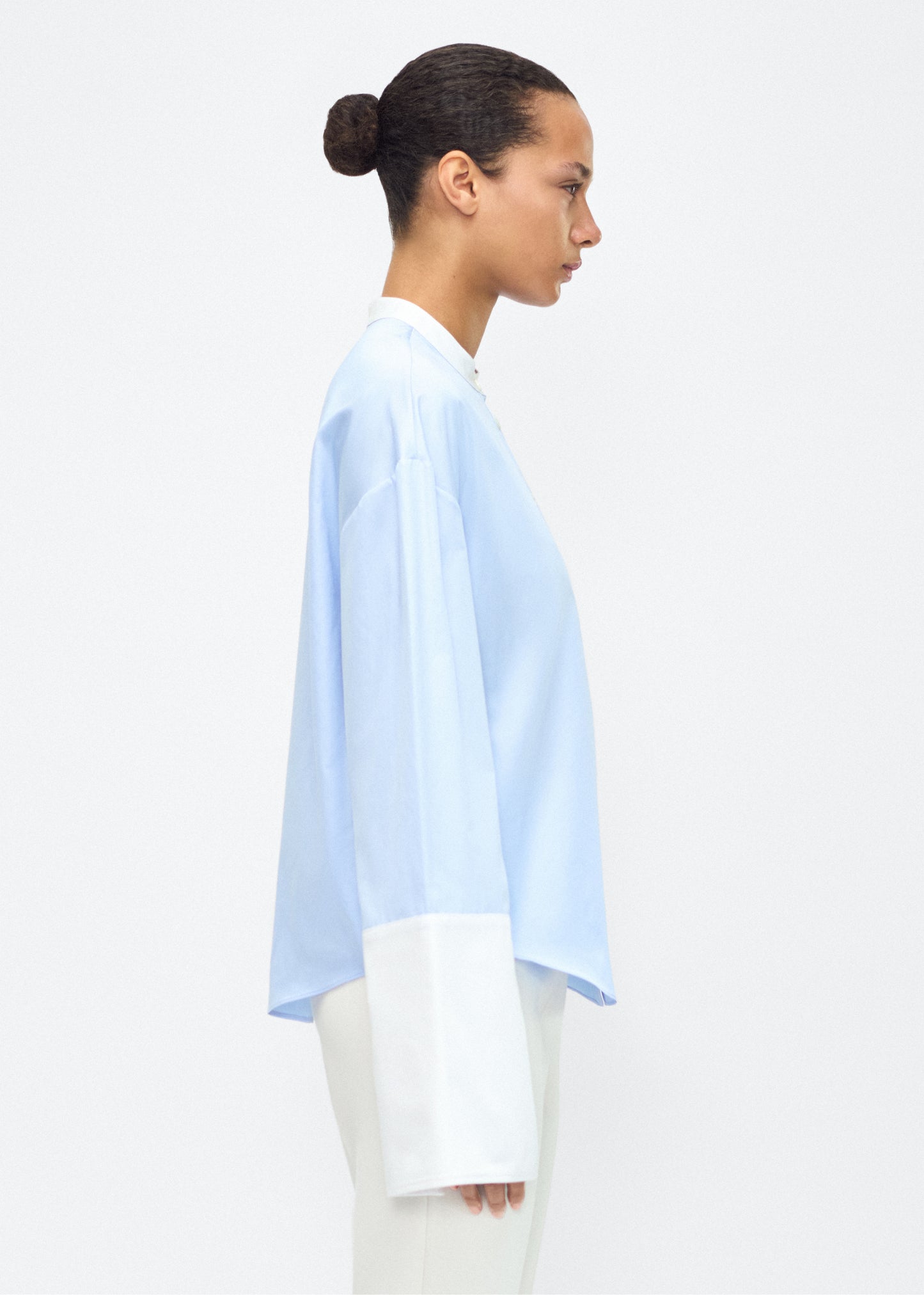 Model facing side – bram top, P25139CS, Pale Blue – Adam Lippes, P25 collection: A new shape from the atelier, the Bram Top is crafted from soft organic cotton poplin in a refreshing pale blue hue. The relaxed fit i enhanced by contrasting white cuffs and collar, with a subtle slit near the cuffs for added detail. Made from the highest quality cotton and featuring diamond monogram mother of pearl buttons down the center front, the top offers elegance and comfort.