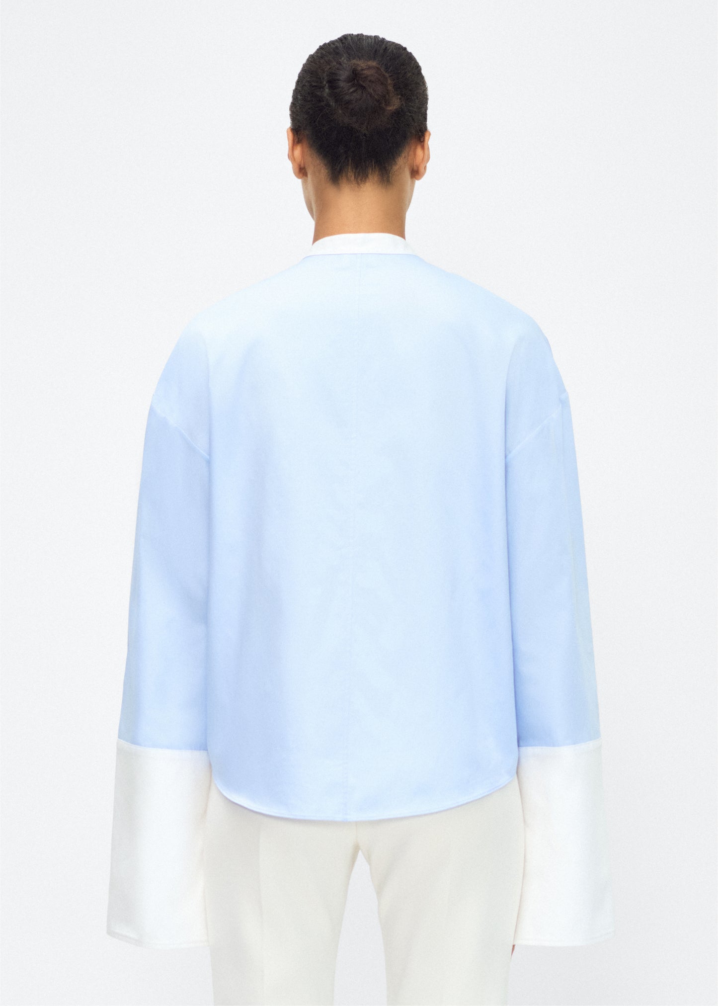 Model facing back – bram top, P25139CS, Pale Blue – Adam Lippes, P25 collection: A new shape from the atelier, the Bram Top is crafted from soft organic cotton poplin in a refreshing pale blue hue. The relaxed fit i enhanced by contrasting white cuffs and collar, with a subtle slit near the cuffs for added detail. Made from the highest quality cotton and featuring diamond monogram mother of pearl buttons down the center front, the top offers elegance and comfort.