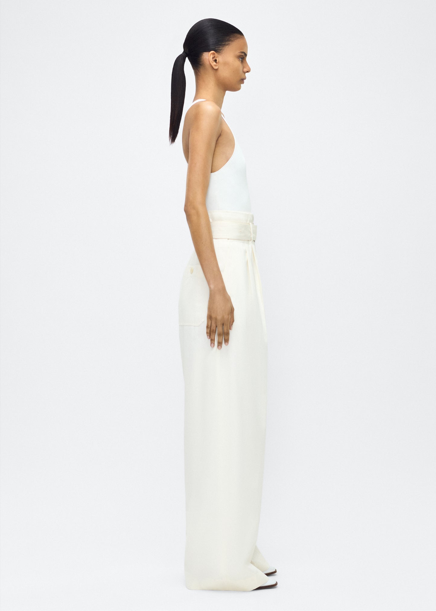 Image of model facing side – Jericho Pant, P25505WI, Ivory – Adam Lippes, P25 collection: The Jericho Pant is crafted from luxurious cotton-cashmere twill, offering the perfect balance of softness and structure. Pleated fronts and a wide-leg design create an easy, relaxed fit, while a belt at the waist adds elegant definition.