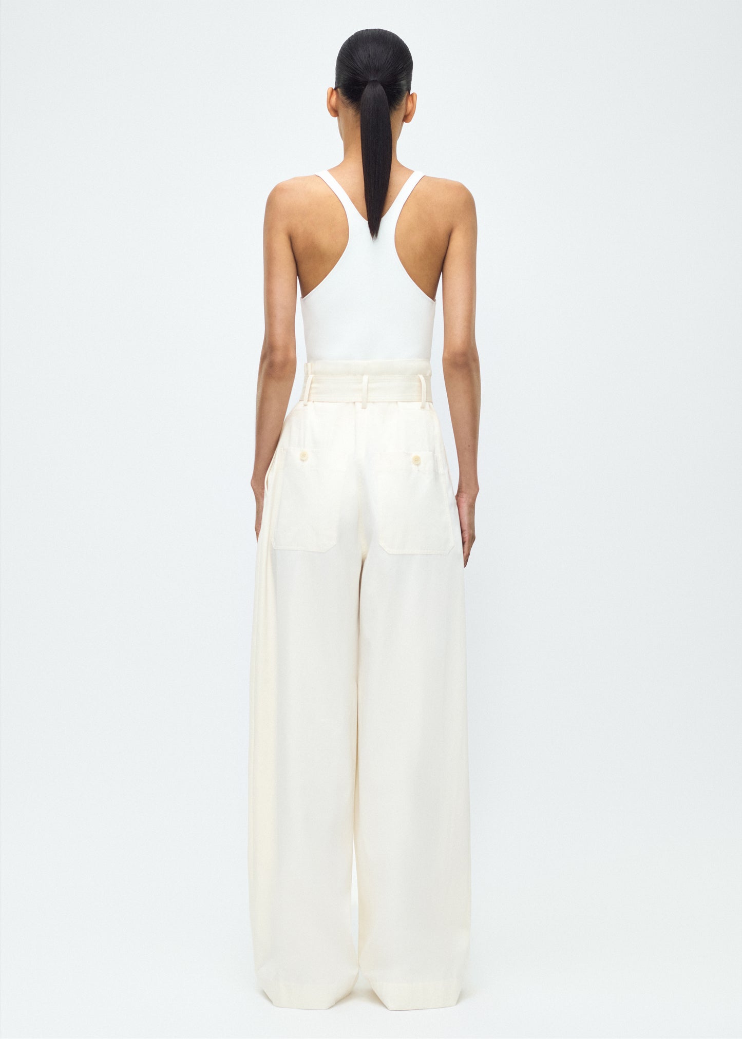 Image of model facing back – Jericho Pant, P25505WI, Ivory – Adam Lippes, P25 collection: The Jericho Pant is crafted from luxurious cotton-cashmere twill, offering the perfect balance of softness and structure. Pleated fronts and a wide-leg design create an easy, relaxed fit, while a belt at the waist adds elegant definition.