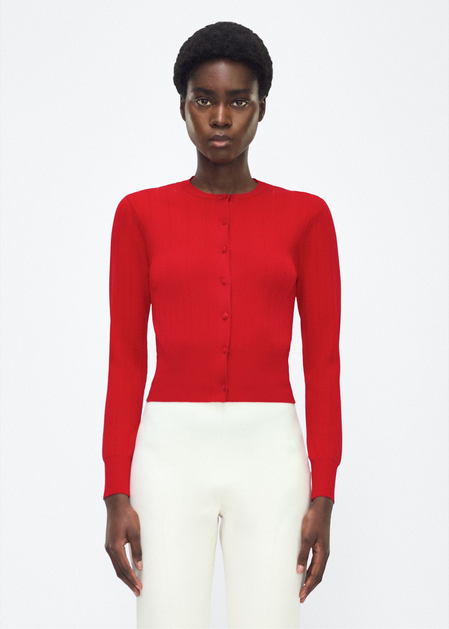 Image of model facing front, P25625UB, Faye Cardigan in Poppy Adam Lippes P25 collection: A classic pointelle stitch cardigan, crafted in our signature compact jacquard. Designed with a fitted bodice and delicate stitching, the piece is finished with covered buttons along the front placket for a refined look.