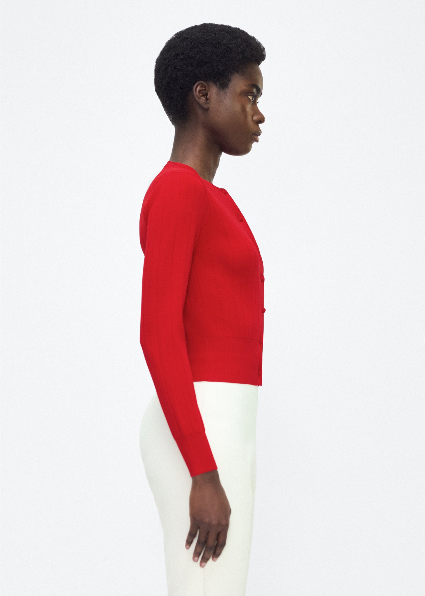 Image of model facing side P25625UB, Faye Cardigan in poppy Adam Lippes P25 collection: A classic pointelle stitch cardigan, crafted in our signature compact jacquard. Designed with a fitted bodice and delicate stitching, the piece is finished with covered buttons along the front placket for a refined look.