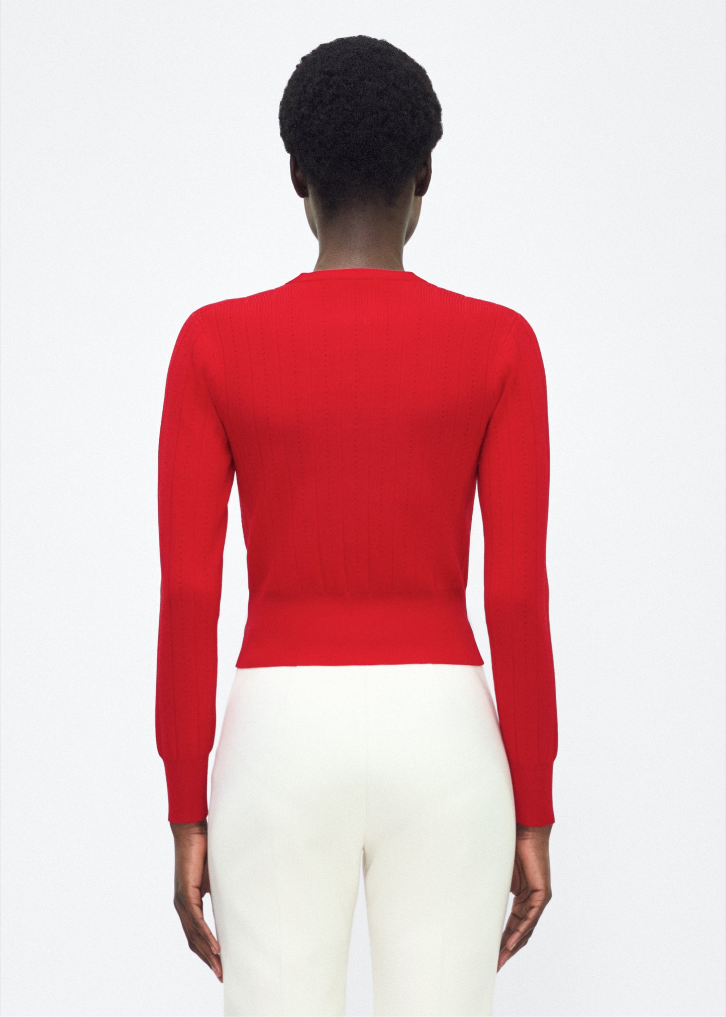 Image of model facing back P25625UB, Faye Cardigan in poppy Adam Lippes P25 collection: A classic pointelle stitch cardigan, crafted in our signature compact jacquard. Designed with a fitted bodice and delicate stitching, the piece is finished with covered buttons along the front placket for a refined look.