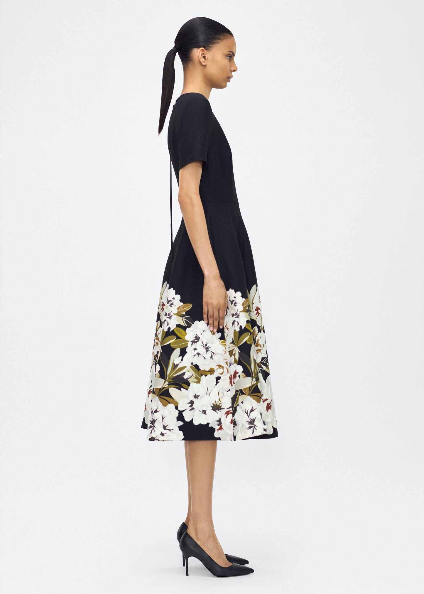 Model facing side, wearing P25722PW, the Evangeline Dress in Black/white; From the Adam Lippes P25 Collection, A signature silhouette from the atelier, the Evangeline Dress is crafted in luxurious printed silk wool adorned with this season’s Sicily-inspired floral motif. Designed with an A-line silhouette and a gently flared skirt, it drapes gracefully for an elegant look.