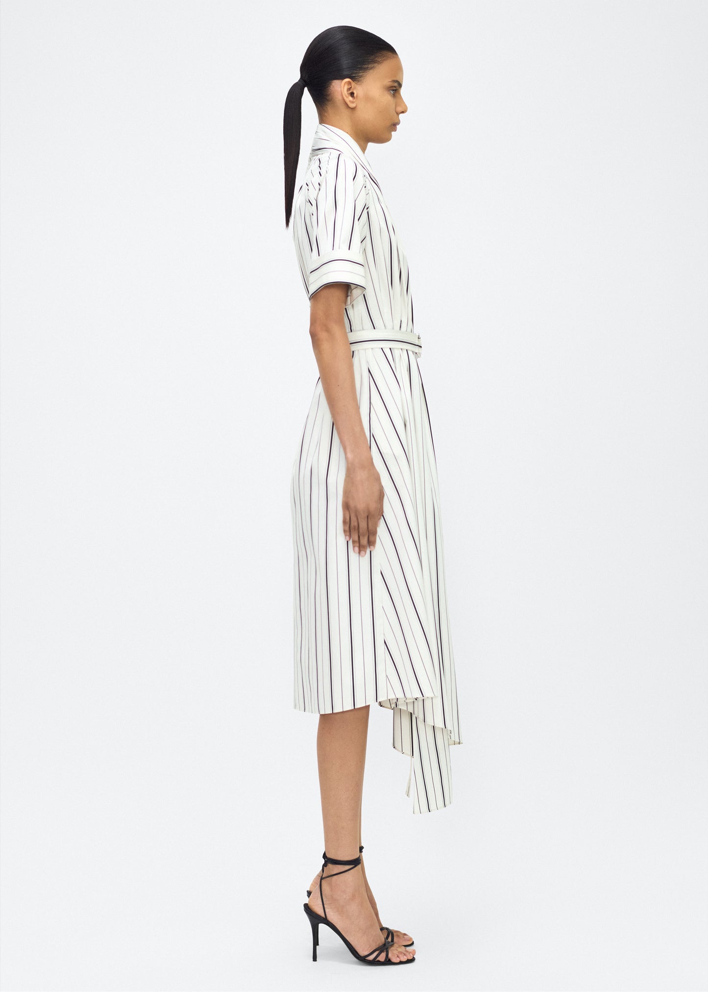 Model facing side, P25723SS, Ivory/Black, P25 Adam Lippes collection: The Nansi Dress is crafted from crisp stripe poplin in ivory and black, featuring an asymmetrical hem that adds a modern twist to a classic design. A sleek V-neckline and short sleeves enhance its effortless sophistication, while a fabric belt cinches the waist for a defined silhouette.