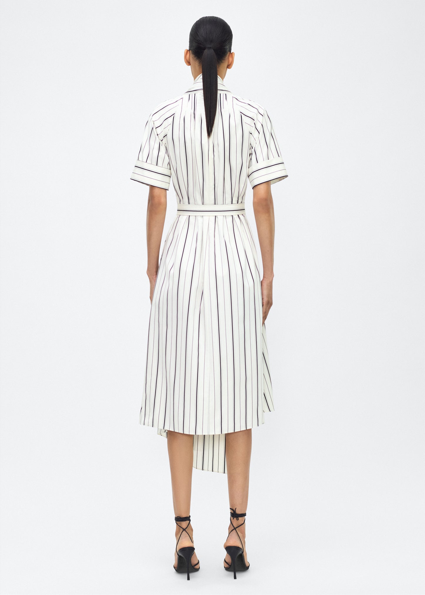 Model facing back, P25723SS, Ivory/Black, P25 Adam Lippes collection: The Nansi Dress is crafted from crisp stripe poplin in ivory and black, featuring an asymmetrical hem that adds a modern twist to a classic design. A sleek V-neckline and short sleeves enhance its effortless sophistication, while a fabric belt cinches the waist for a defined silhouette.