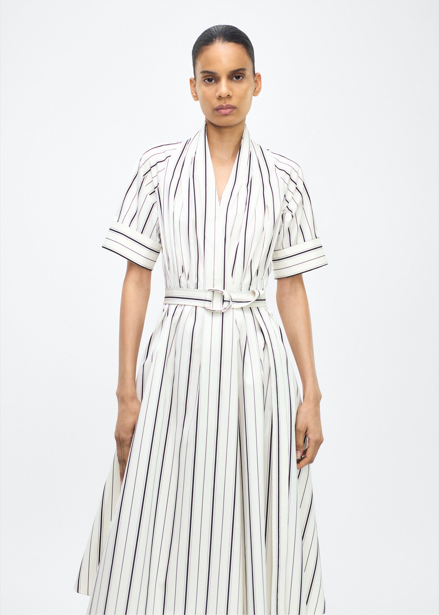 Model facing front, P25723SS, Ivory/Black, P25 Adam Lippes collection: The Nansi Dress is crafted from crisp stripe poplin in ivory and black, featuring an asymmetrical hem that adds a modern twist to a classic design. A sleek V-neckline and short sleeves enhance its effortless sophistication, while a fabric belt cinches the waist for a defined silhouette.
