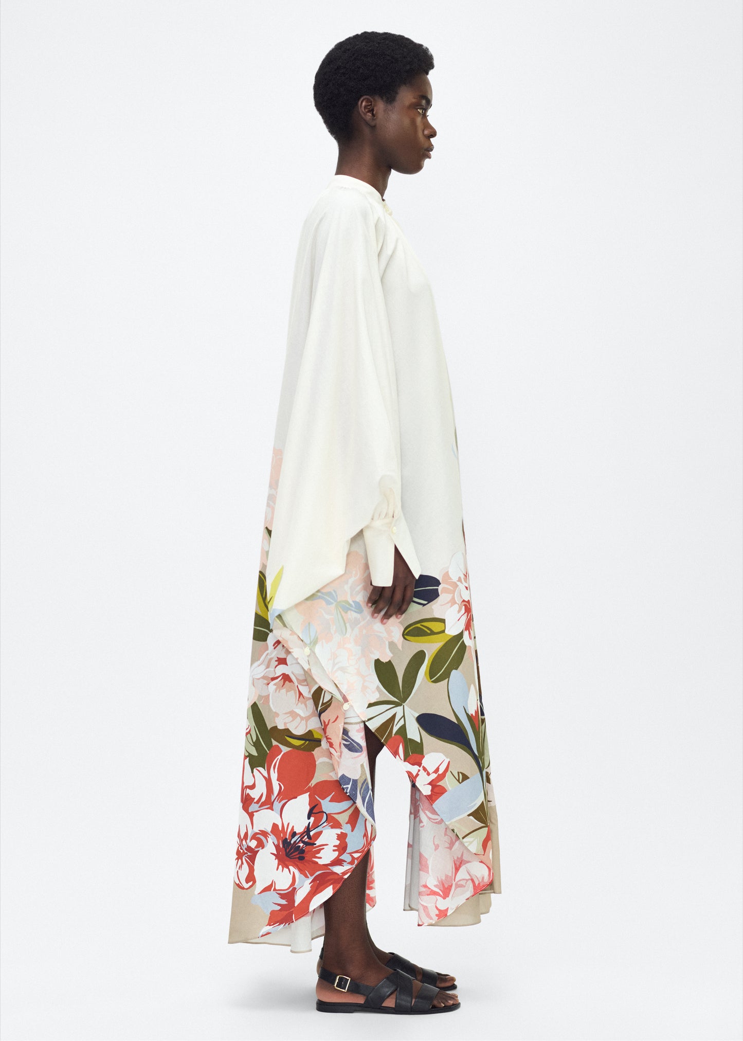 Model facing side, P25726PV, Rysa Caftan, Champagne/Multi, Adam Lippes P25 Collection; A new shape from the atelier, the Rysa Caftan is crafted from airy printed voile, featuring this season’s Sicily-inspired floral motif. The fluid hem adds a modern twist to the traditional caftan silhouette. With its flowing, relaxed fit, it exudes effortless elegance. Features a button loop closure and diamond monogram mother of pearl buttons.