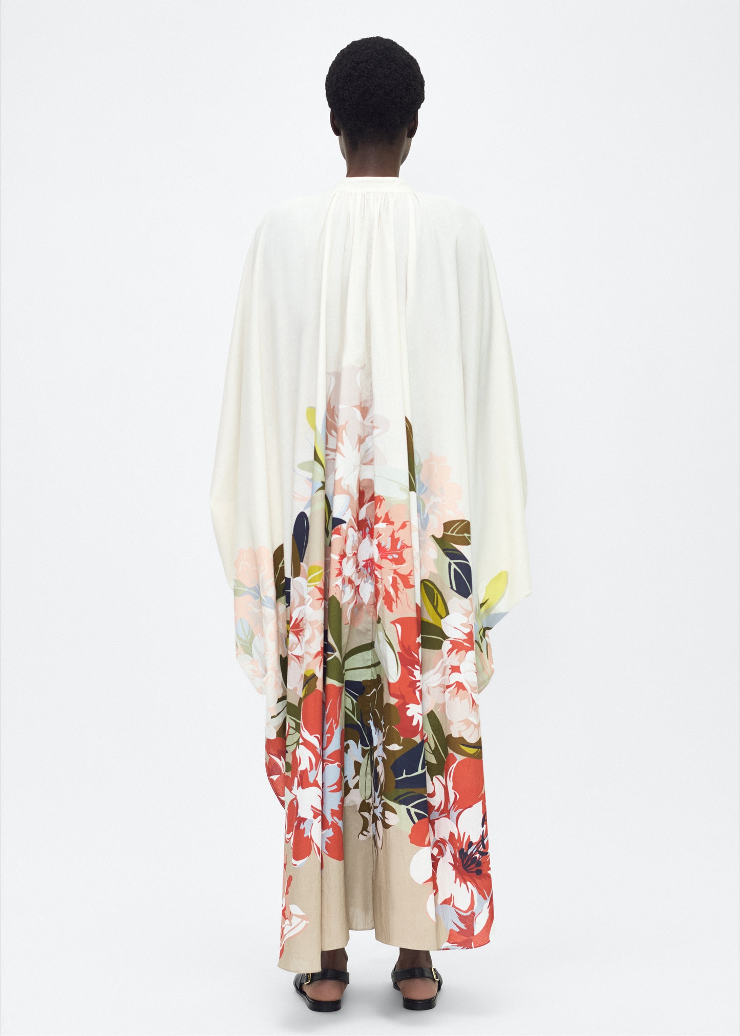 Model facing back, P25726PV, Rysa Caftan, Champagne/Multi, Adam Lippes P25 Collection; A new shape from the atelier, the Rysa Caftan is crafted from airy printed voile, featuring this season’s Sicily-inspired floral motif. The fluid hem adds a modern twist to the traditional caftan silhouette. With its flowing, relaxed fit, it exudes effortless elegance. Features a button loop closure and diamond monogram mother of pearl buttons.