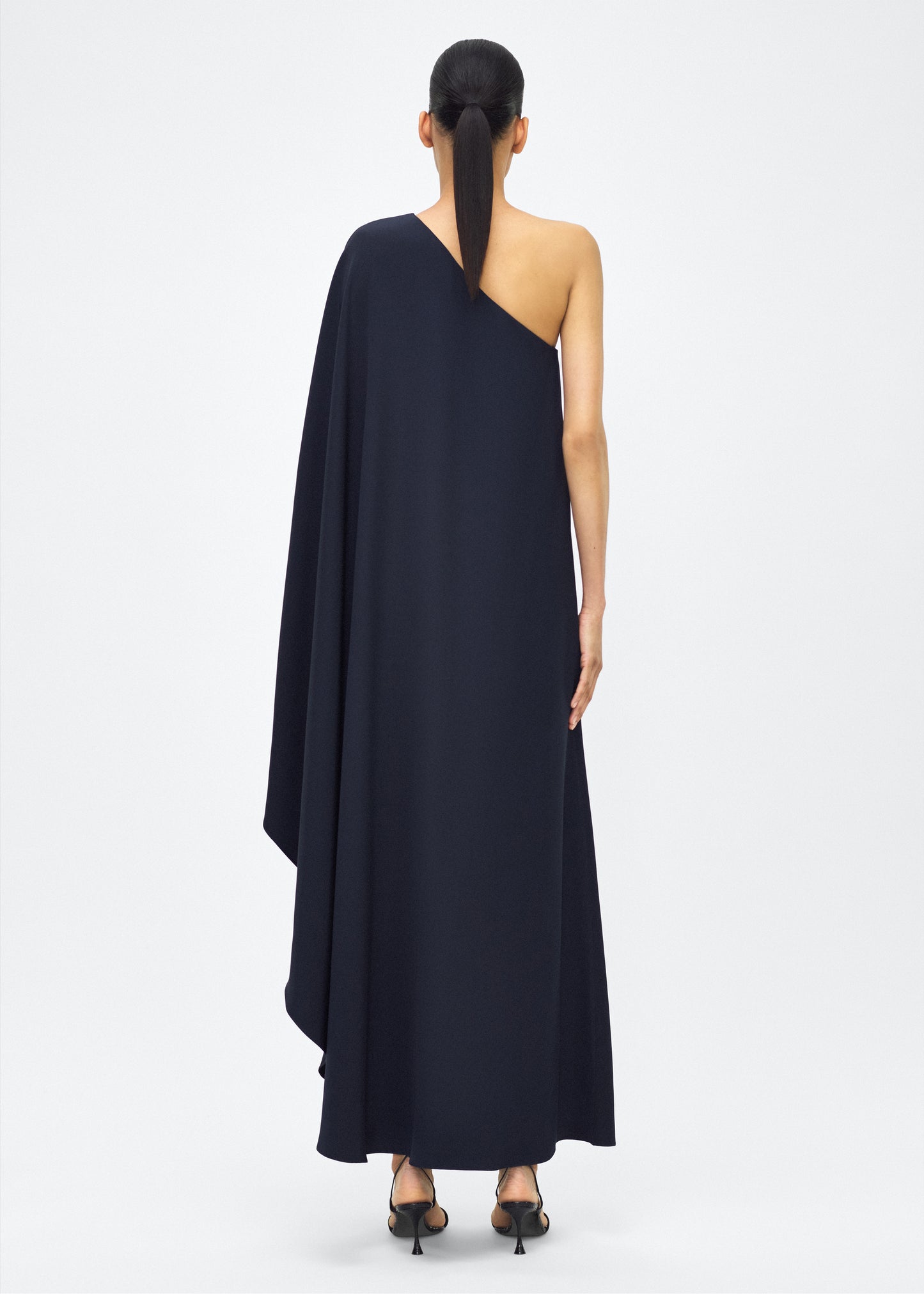 Image of Model facing back, P25747SE, Navy, Adam Lippes P25 Collection; The Solene Dress is a striking one-shoulder design crafted from luxurious silk crepe. The fabric drapes gracefully, creating an elegant, fluid silhouette. The asymmetrical cut is complemented by the soft, flowing lines that cascade effortlessly.