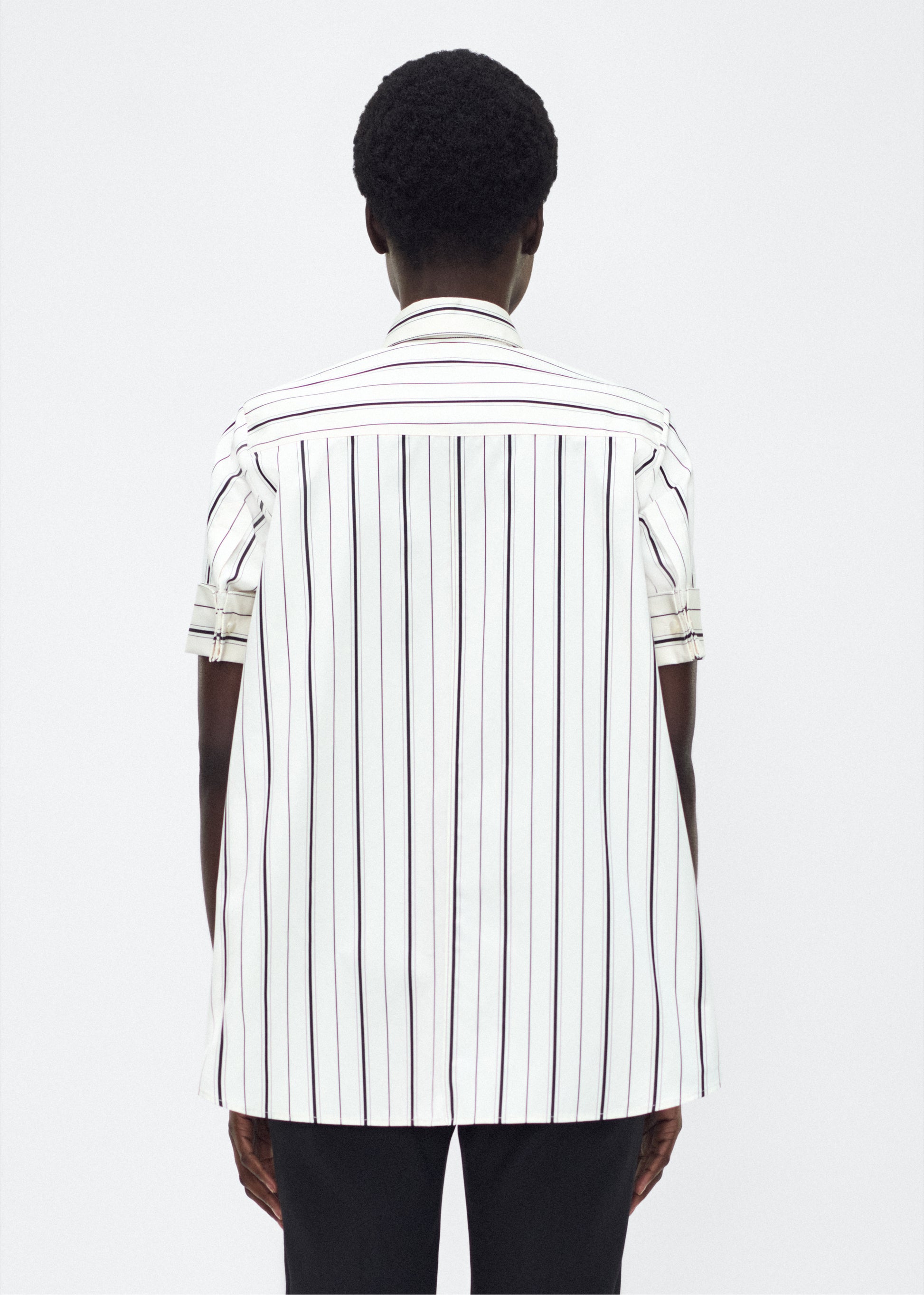 Model back facing image – Trapeze Shirt, P25106SS, Ivory/Black – Adam Lippes, P25 collection: The Adam Lippes Trapeze Shirt remains a timeless favorite from the atelier. Crafted from crisp Italian cotton, this season’s version features a classic ivory and black stripe. The piece features diamond monogram mother of pearl buttons along the center front placket, collar stand and French cuffs. Designed with side slits for a breezy, relaxed fit.