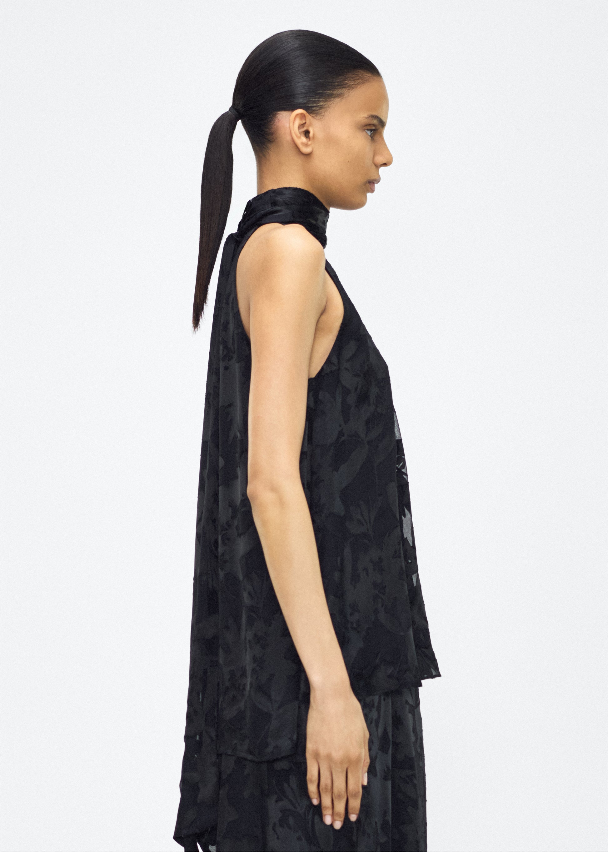 Image of model facing side – giulia top, P25117DV, Black – Adam Lippes, P25 collection: The Giulia Top is crafted from elegant burn-out fabric, and features an intricate floral design. The classic black hue enhances the rich texture, while the flowing silhouette exudes sophistication. Pair with the Emi Skirt in Devore for a coordinated ensemble.