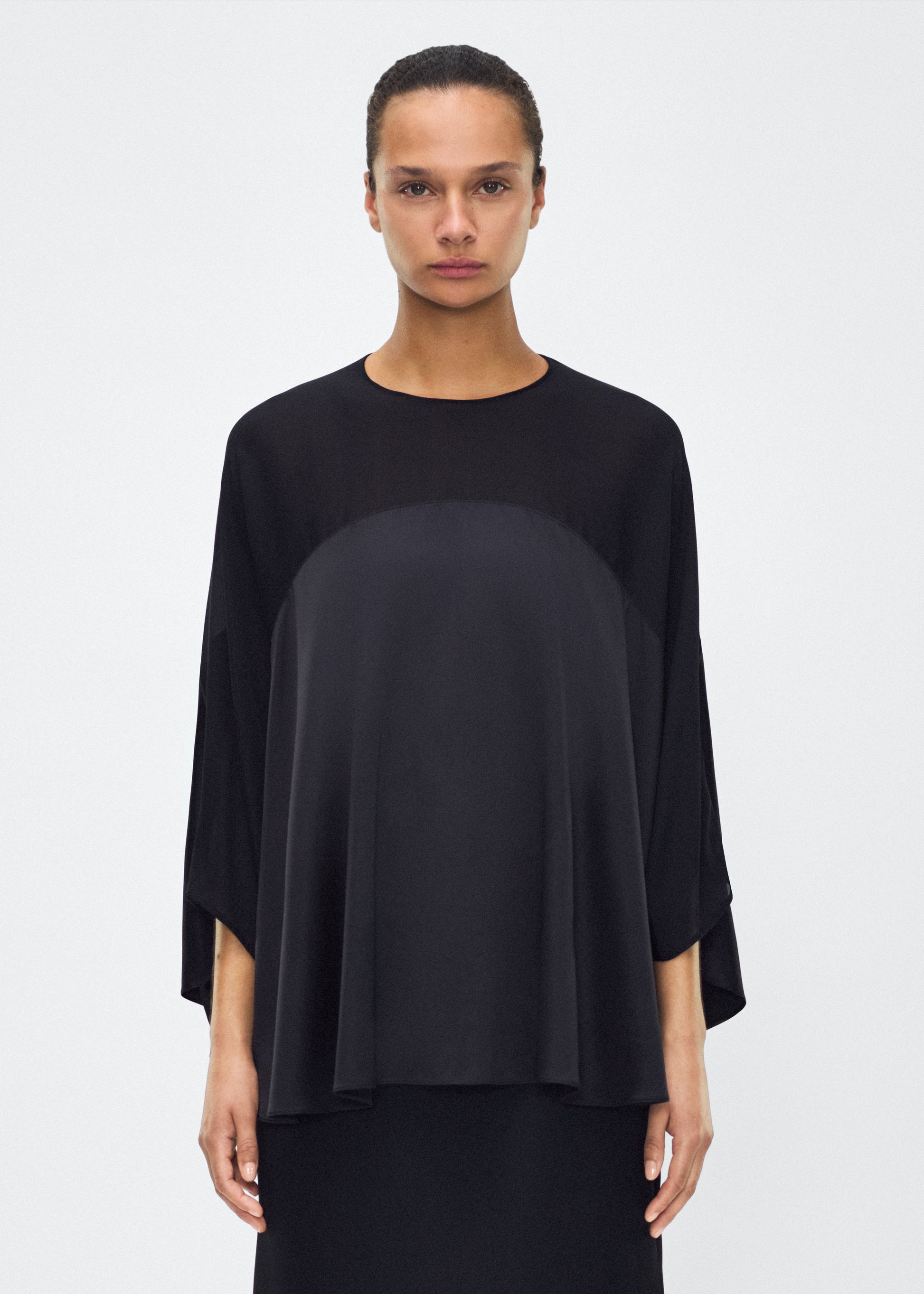 Image of model facing forward– apollo top, P25127DC, black – Adam Lippes, P25 collection: A new shape from the atelier, the Apollo Top is crafted from luxurious 100% silk in a timeless black hue. The flowing, draped design features a high-neck collar and a cascading silhouette, exuding modern elegance. 