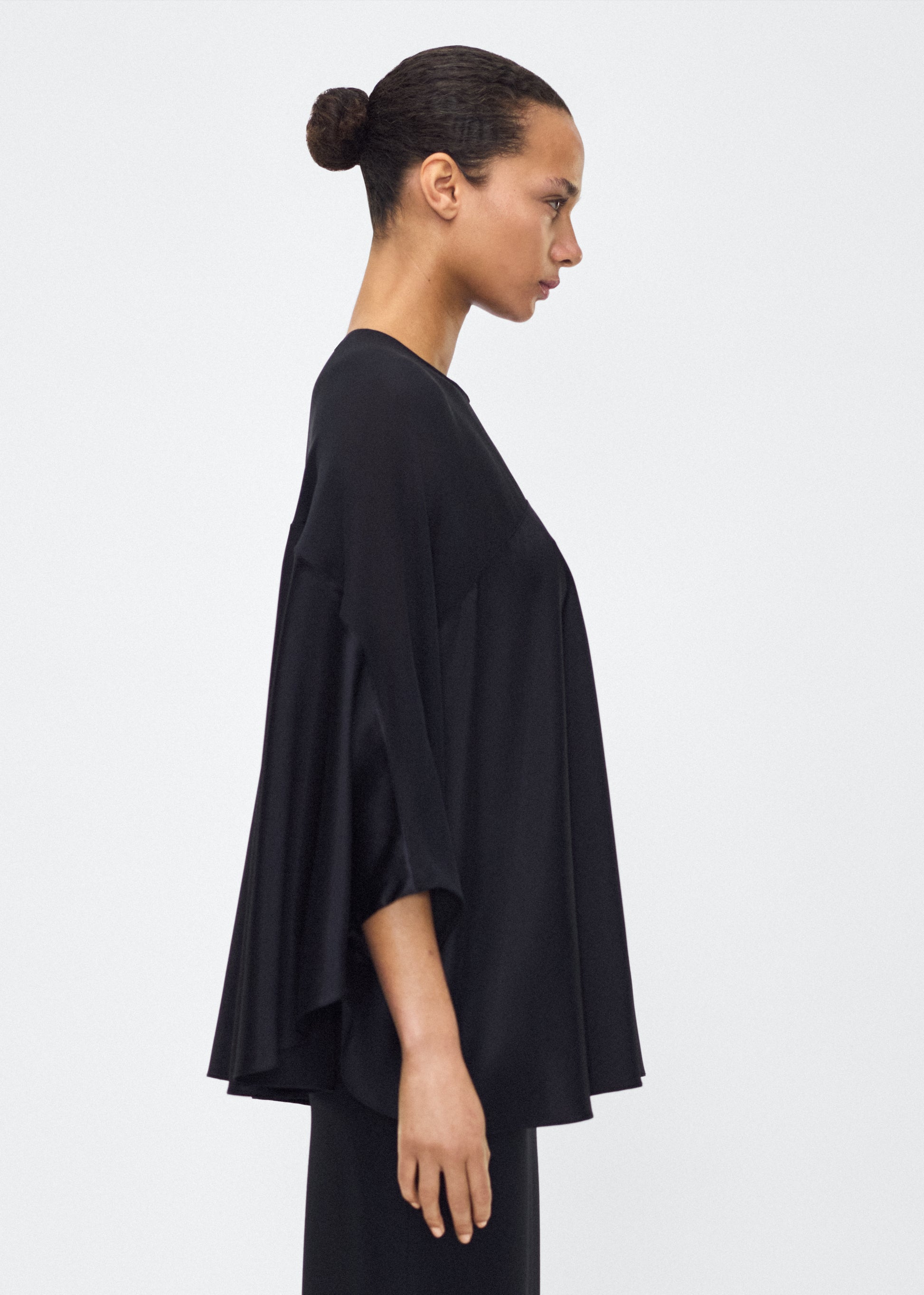 Image of model facing side wearing the apollo top, P25127DC, black – Adam Lippes, P25 collection: A new shape from the atelier, the Apollo Top is crafted from luxurious 100% silk in a timeless black hue. The flowing, draped design features a high-neck collar and a cascading silhouette, exuding modern elegance. 