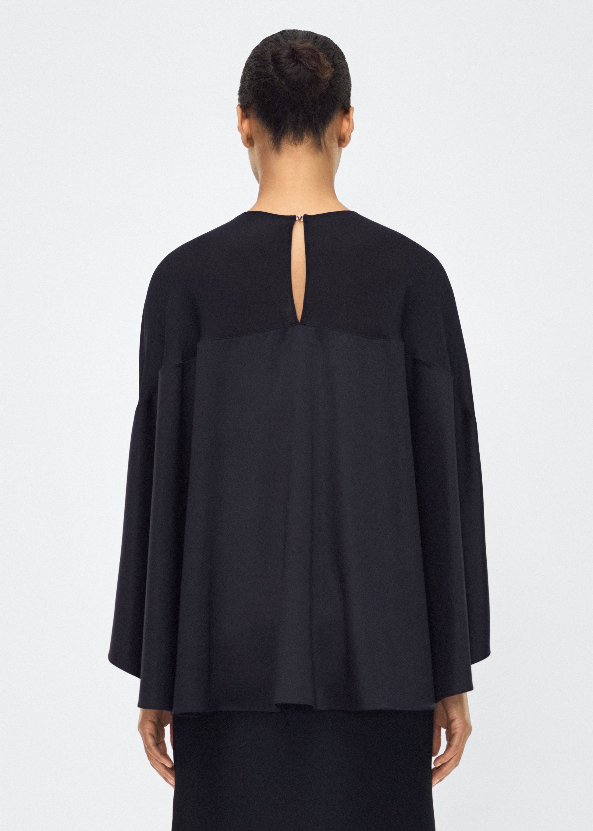 Image of model facing back wearing the apollo top, P25127DC, black – Adam Lippes, P25 collection: A new shape from the atelier, the Apollo Top is crafted from luxurious 100% silk in a timeless black hue. The flowing, draped design features a high-neck collar and a cascading silhouette, exuding modern elegance. 