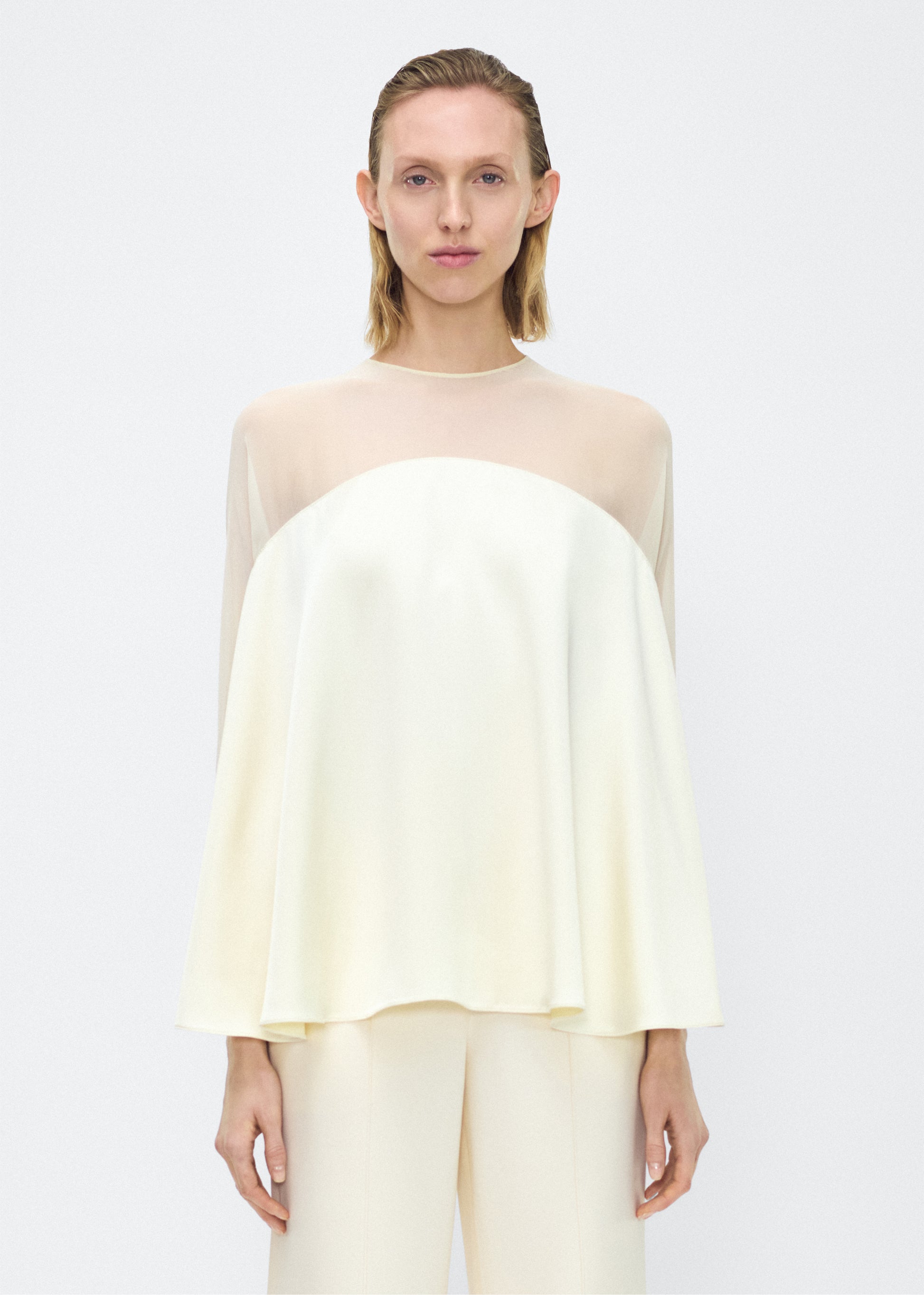 Model facing front – apollo top, P25127DC, cream – Adam Lippes, P25 collection: A new shape from the atelier, the Apollo Top is crafted from luxurious 100% silk in a soft cream hue. The flowing, draped design features a high-neck collar and a cascading silhouette, exuding modern elegance. 