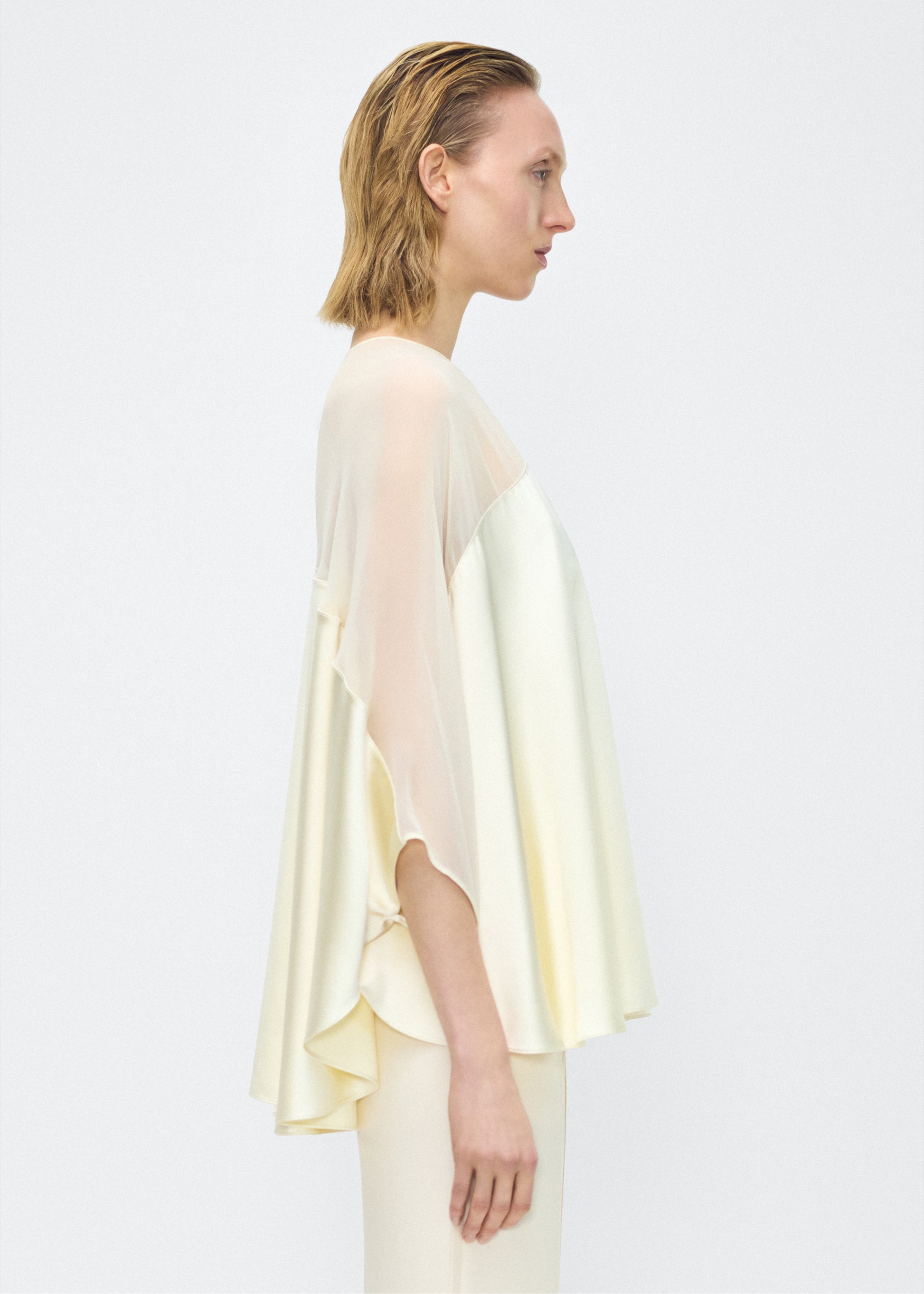 Model facing side – apollo top, P25127DC, cream – Adam Lippes, P25 collection: A new shape from the atelier, the Apollo Top is crafted from luxurious 100% silk in a soft cream hue. The flowing, draped design features a high-neck collar and a cascading silhouette, exuding modern elegance. 