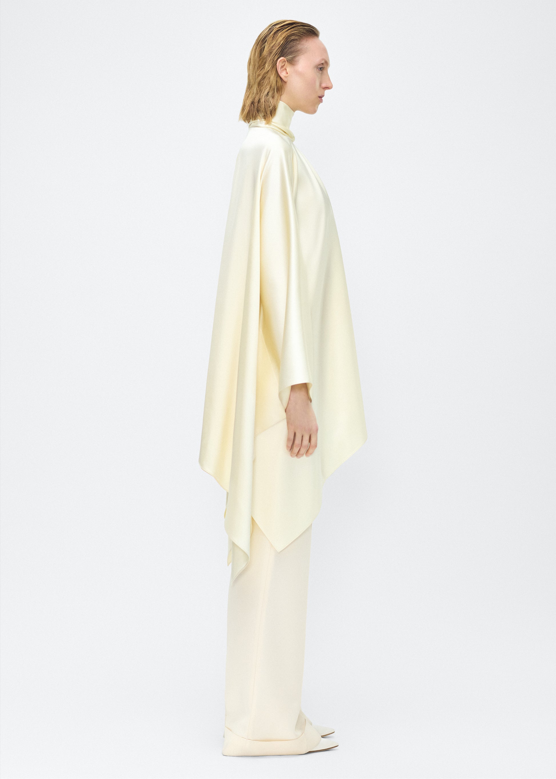 Model facing side – tana top , P25134HM, cream – Adam Lippes, P25 collection: The Tana Top is crafted from luxurious double-face charmeuse, offering a rich, fluid drape in a delicate cream hue. Featuring a detachable scarf for versatile styling options. The clean lines and elegant shape make it a statement piece, perfect for both dressing up and down.
