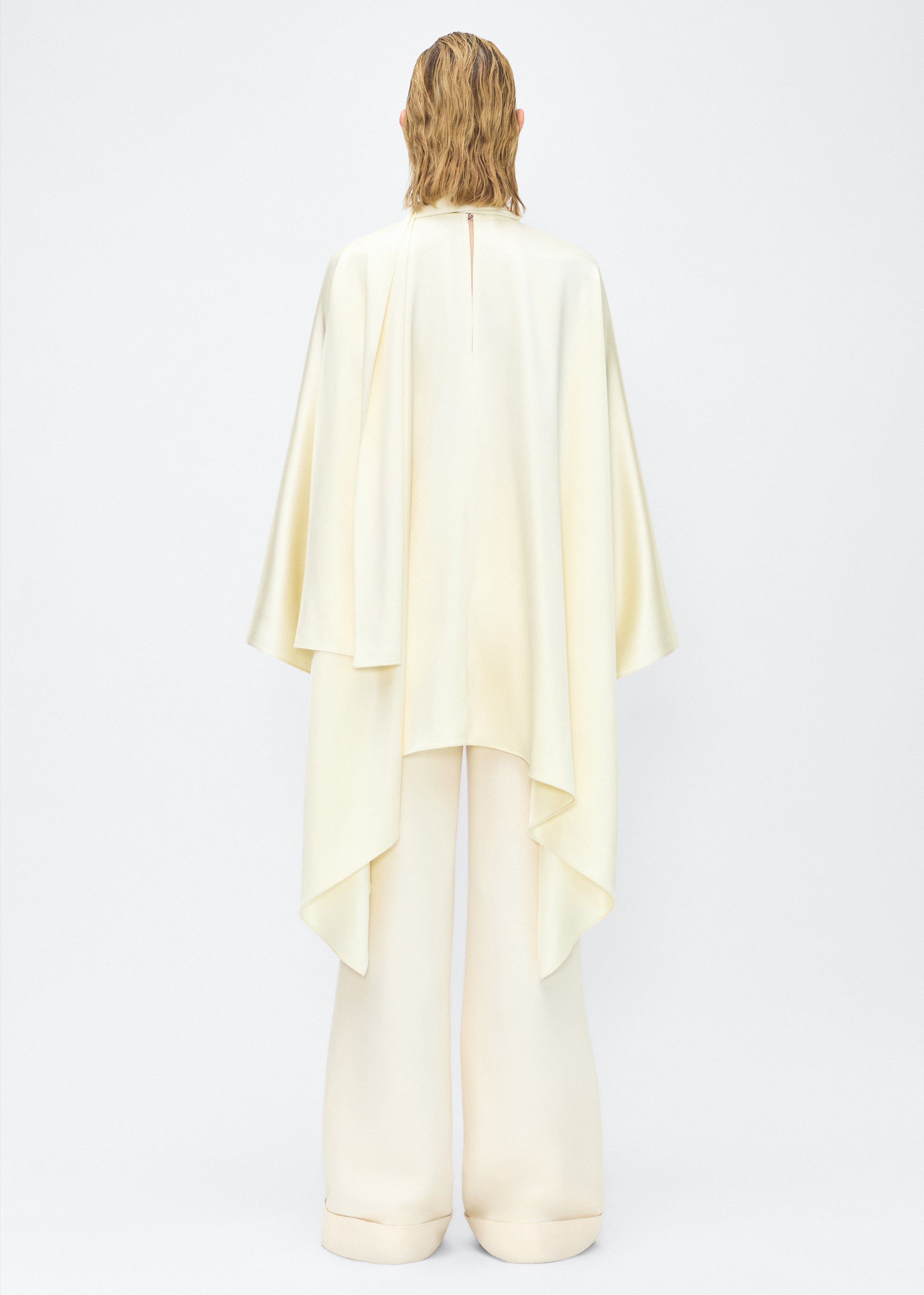Model facing back – tana top , P25134HM, cream – Adam Lippes, P25 collection: The Tana Top is crafted from luxurious double-face charmeuse, offering a rich, fluid drape in a delicate cream hue. Featuring a detachable scarf for versatile styling options. The clean lines and elegant shape make it a statement piece, perfect for both dressing up and down.
