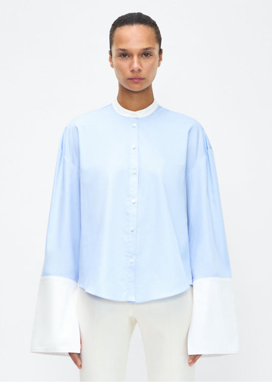 Model facing front – bram top, P25139CS, Pale Blue – Adam Lippes, P25 collection: A new shape from the atelier, the Bram Top is crafted from soft organic cotton poplin in a refreshing pale blue hue. The relaxed fit i enhanced by contrasting white cuffs and collar, with a subtle slit near the cuffs for added detail. Made from the highest quality cotton and featuring diamond monogram mother of pearl buttons down the center front, the top offers elegance and comfort.