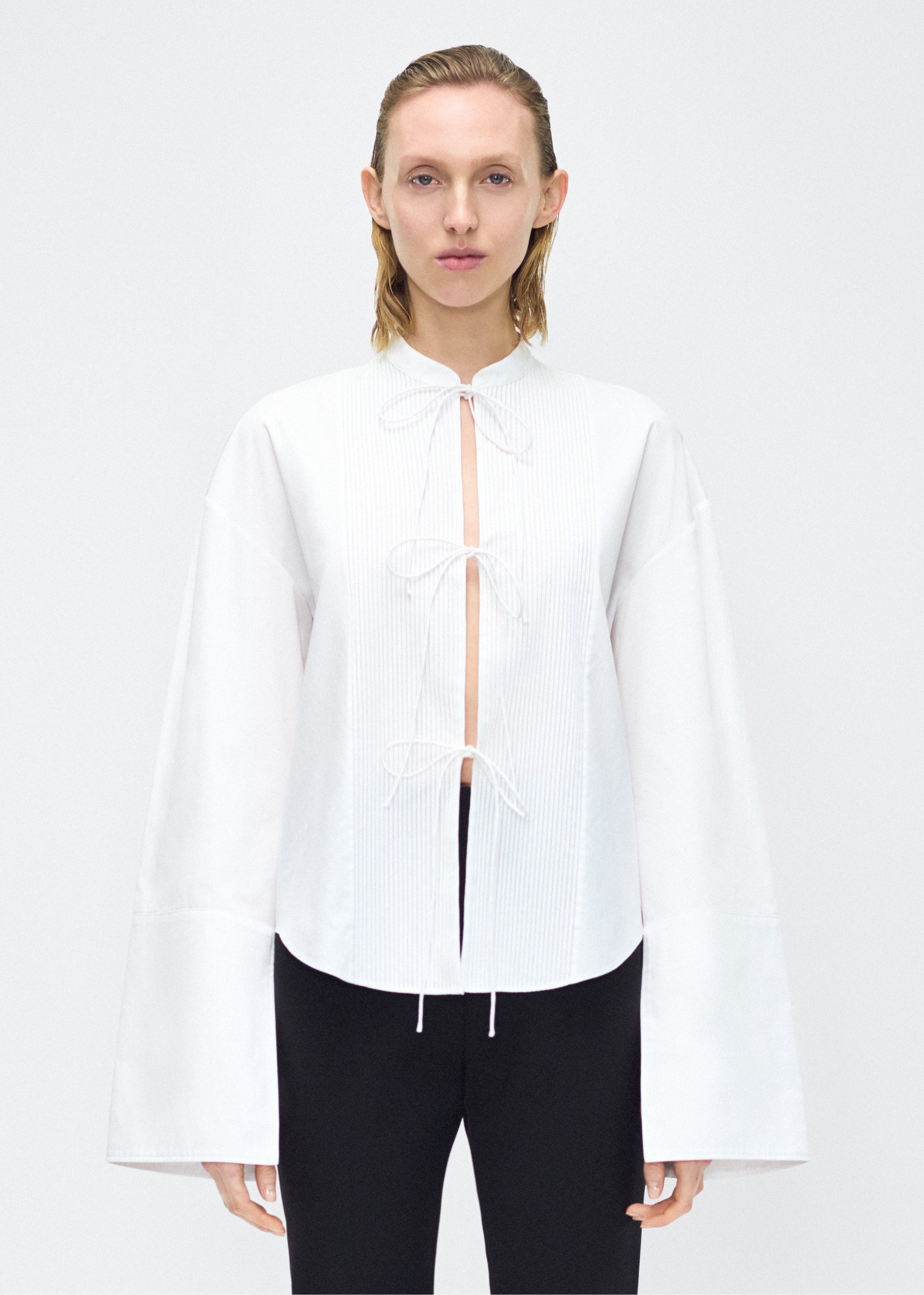 Model facing front – mick top, P25145CS, white – Adam Lippes, P25 collection: A new shape for the season, the Mick Top is crafted from crisp organic cotton poplin in classic white. The top features a tie closure at the center front, adding a subtle touch of modern sophistication. Billowy sleeves with split cuffs create an airy and elegant silhouette. Includes a silk cami for added comfort and versatility.