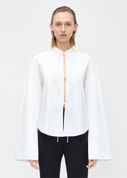 Model facing front – mick top, P25145CS, white – Adam Lippes, P25 collection: A new shape for the season, the Mick Top is crafted from crisp organic cotton poplin in classic white. The top features a tie closure at the center front, adding a subtle touch of modern sophistication. Billowy sleeves with split cuffs create an airy and elegant silhouette. Includes a silk cami for added comfort and versatility.