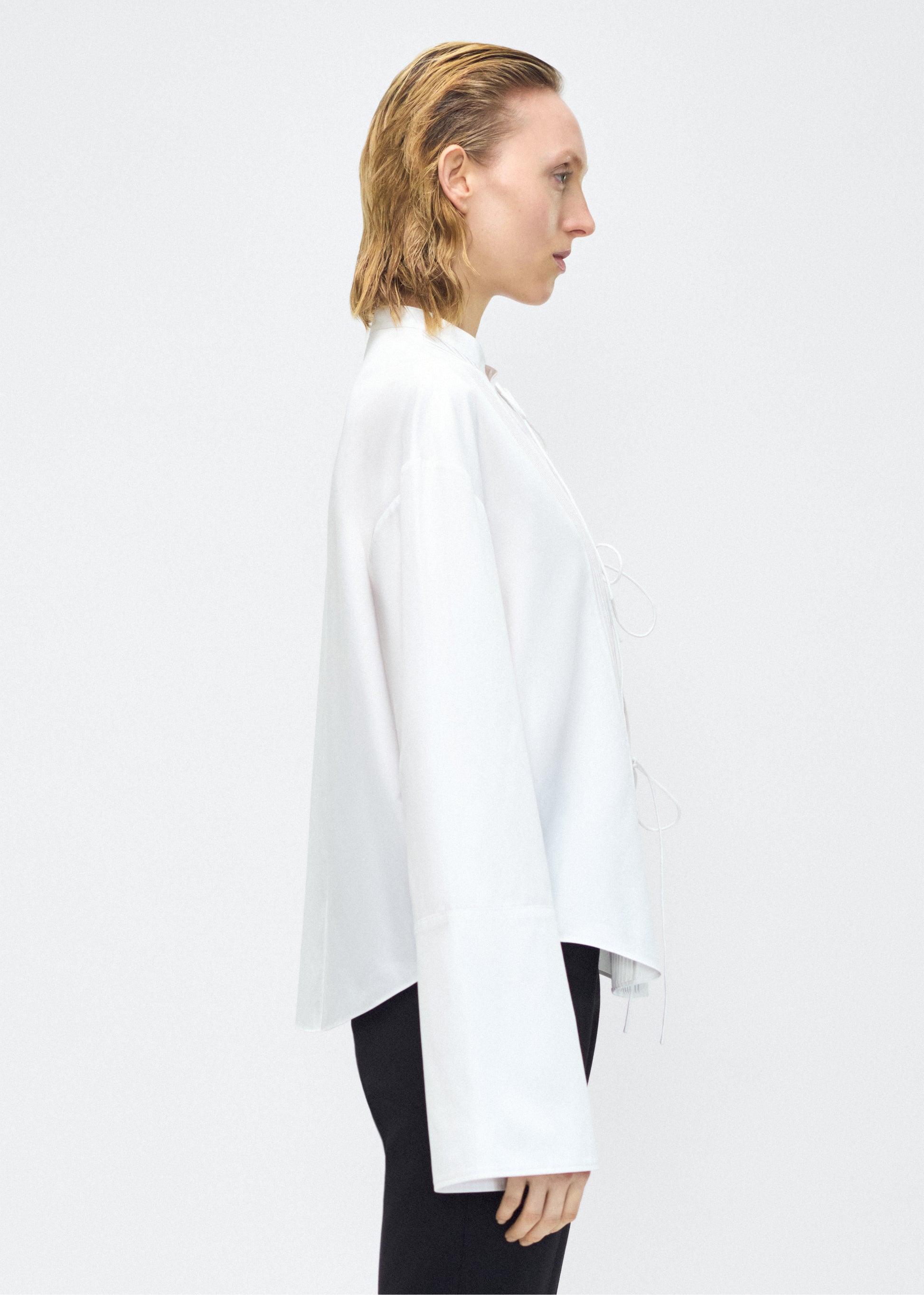 Model facing side – mick top, P25145CS, white – Adam Lippes, P25 collection: A new shape for the season, the Mick Top is crafted from crisp organic cotton poplin in classic white. The top features a tie closure at the center front, adding a subtle touch of modern sophistication. Billowy sleeves with split cuffs create an airy and elegant silhouette. Includes a silk cami for added comfort and versatility.