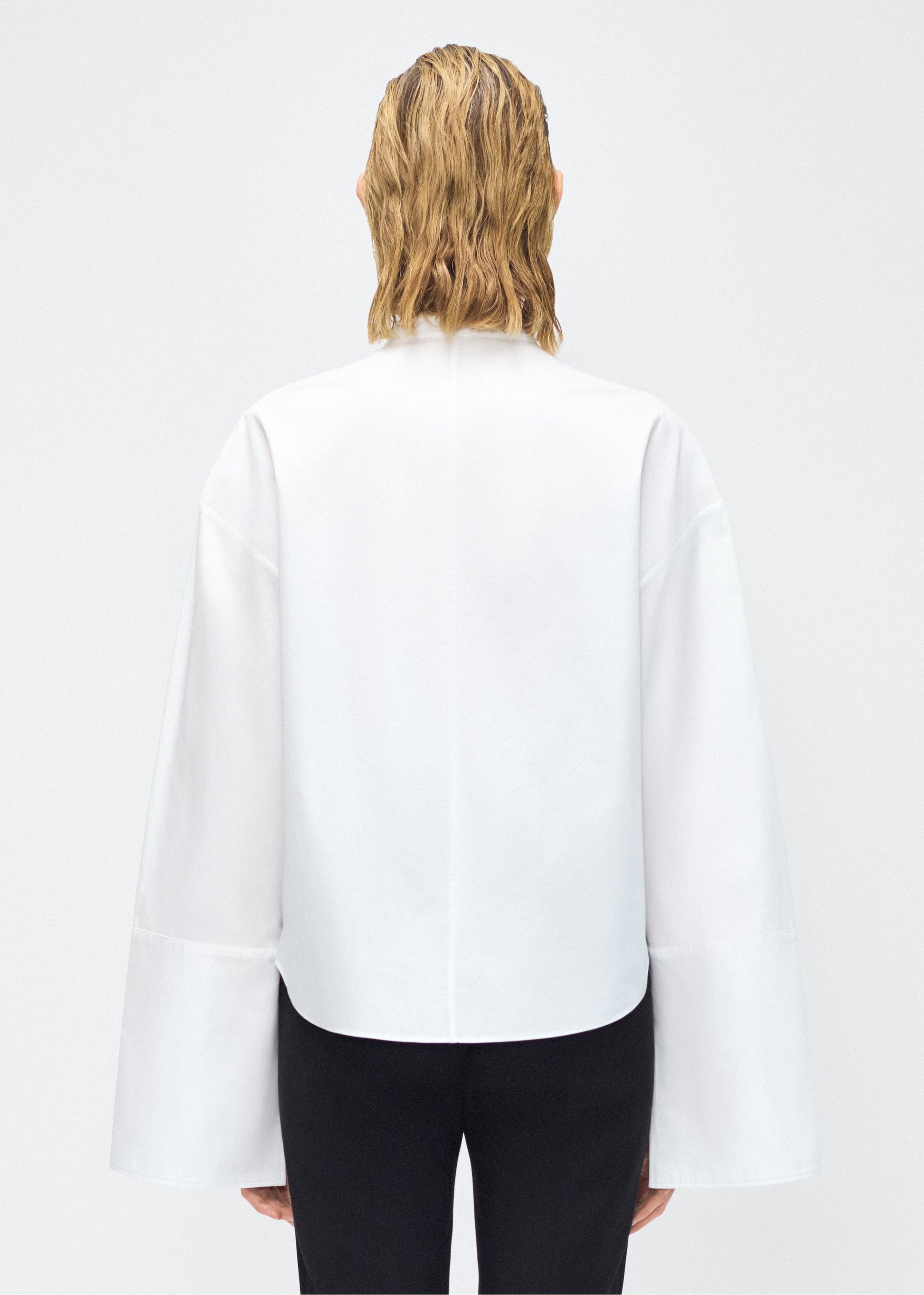 Model facing back – mick top, P25145CS, white – Adam Lippes, P25 collection: A new shape for the season, the Mick Top is crafted from crisp organic cotton poplin in classic white. The top features a tie closure at the center front, adding a subtle touch of modern sophistication. Billowy sleeves with split cuffs create an airy and elegant silhouette. Includes a silk cami for added comfort and versatility.