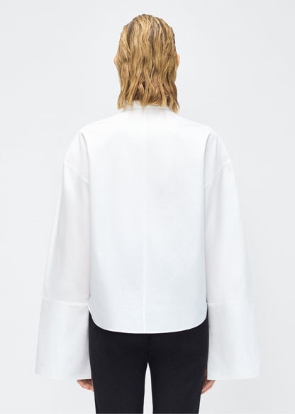 Model facing back – mick top, P25145CS, white – Adam Lippes, P25 collection: A new shape for the season, the Mick Top is crafted from crisp organic cotton poplin in classic white. The top features a tie closure at the center front, adding a subtle touch of modern sophistication. Billowy sleeves with split cuffs create an airy and elegant silhouette. Includes a silk cami for added comfort and versatility.
