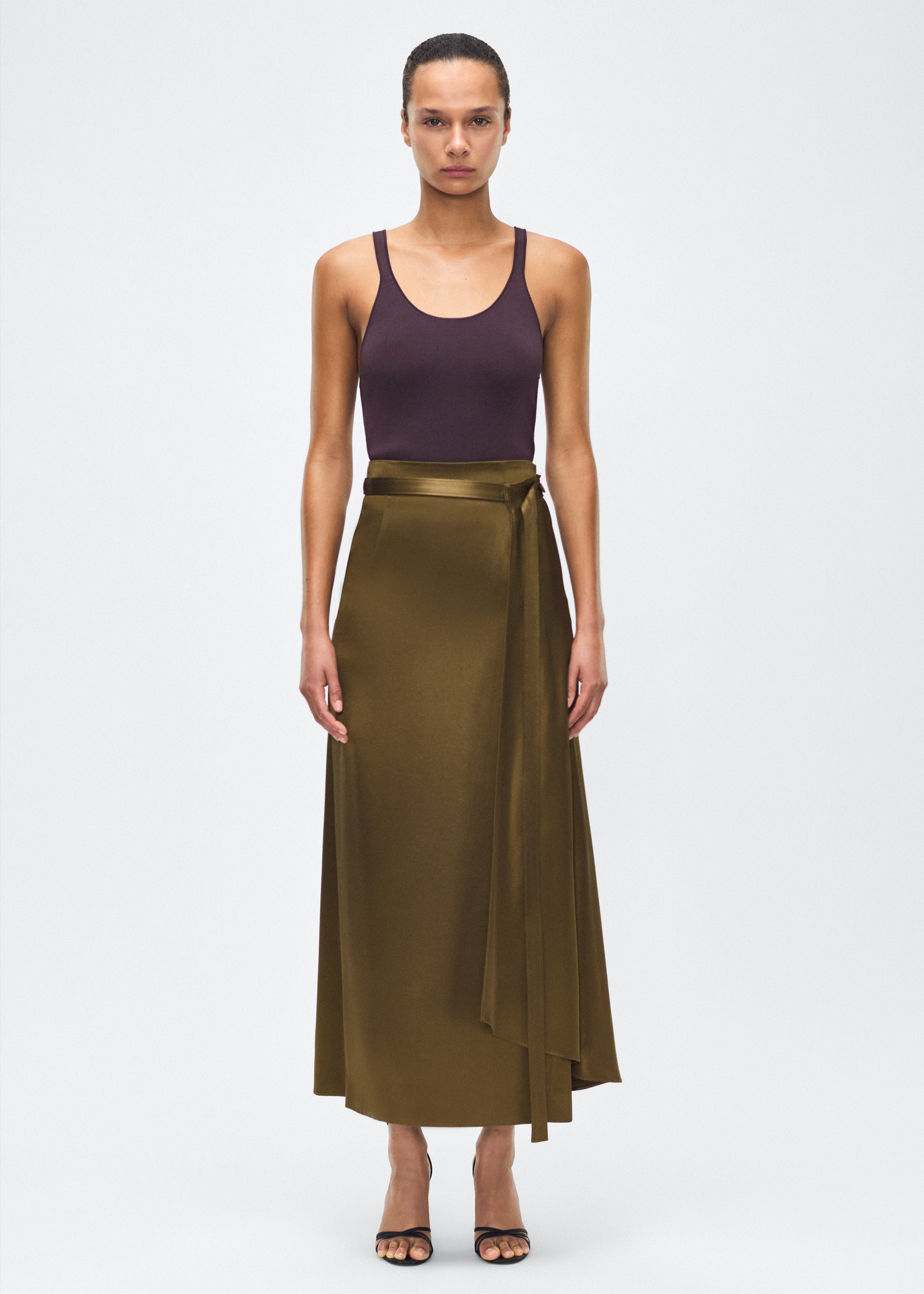 model image front – bruna skirt, P25403HM, olive – Adam Lippes, P25 collection: The Bruna Skirt is crafted from luxurious double-face silk charmeuse, offering a rich sheen and fluid drape. Rendered in a deep olive hue, cascading fabric at the side of the piece adds subtle movement to the elegant silhouette. 
