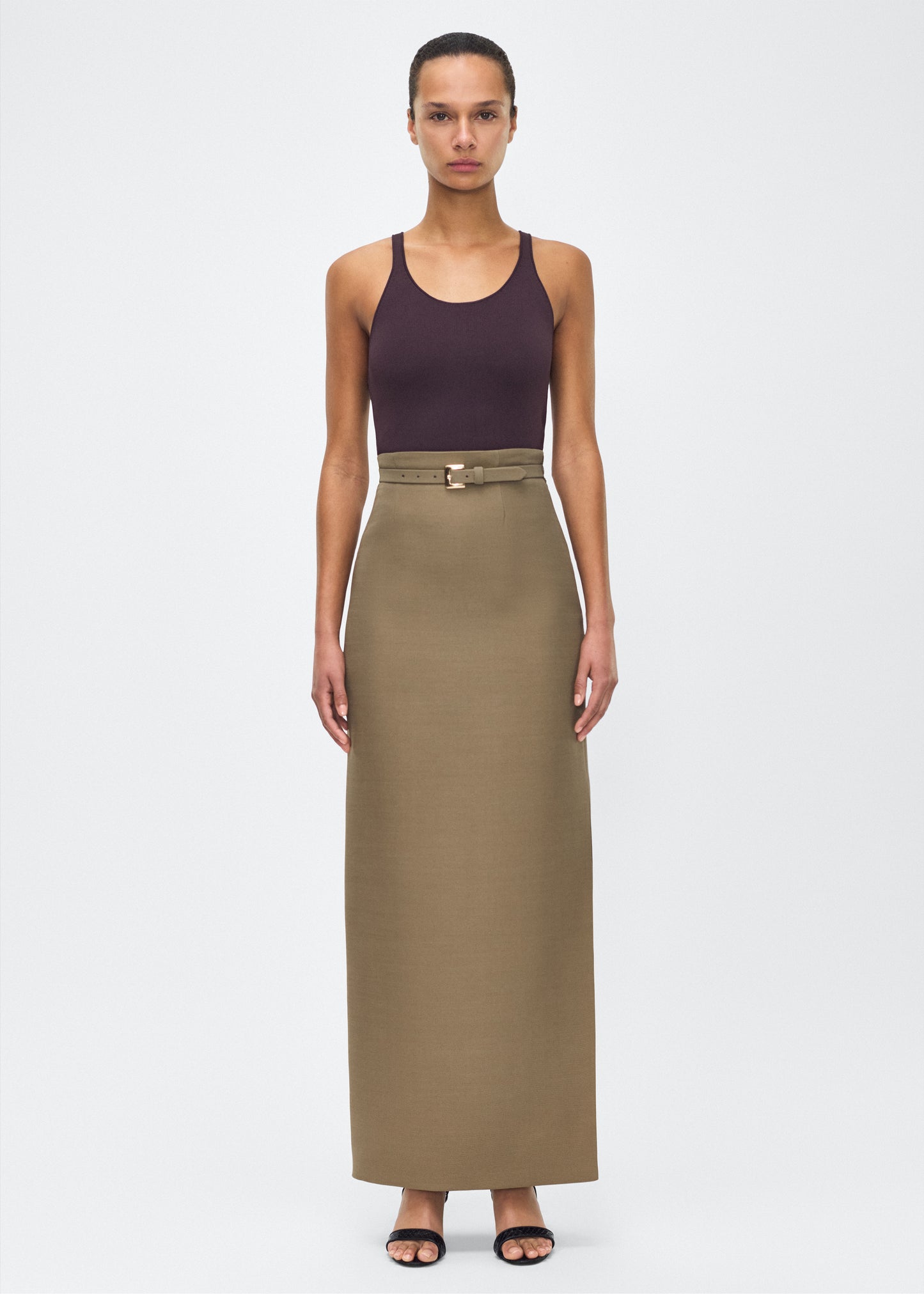 Model facing front wearing Leto Skirt, P25412SW, Military Green – Adam Lippes, P25 collection:The Leto Skirt offers an elevated pencil silhouette, crafted from luxurious silk wool in a sophisticated military green hue. The sleek design is accentuated by a fabric-covered belt, cinching the waist for a tailored fit.