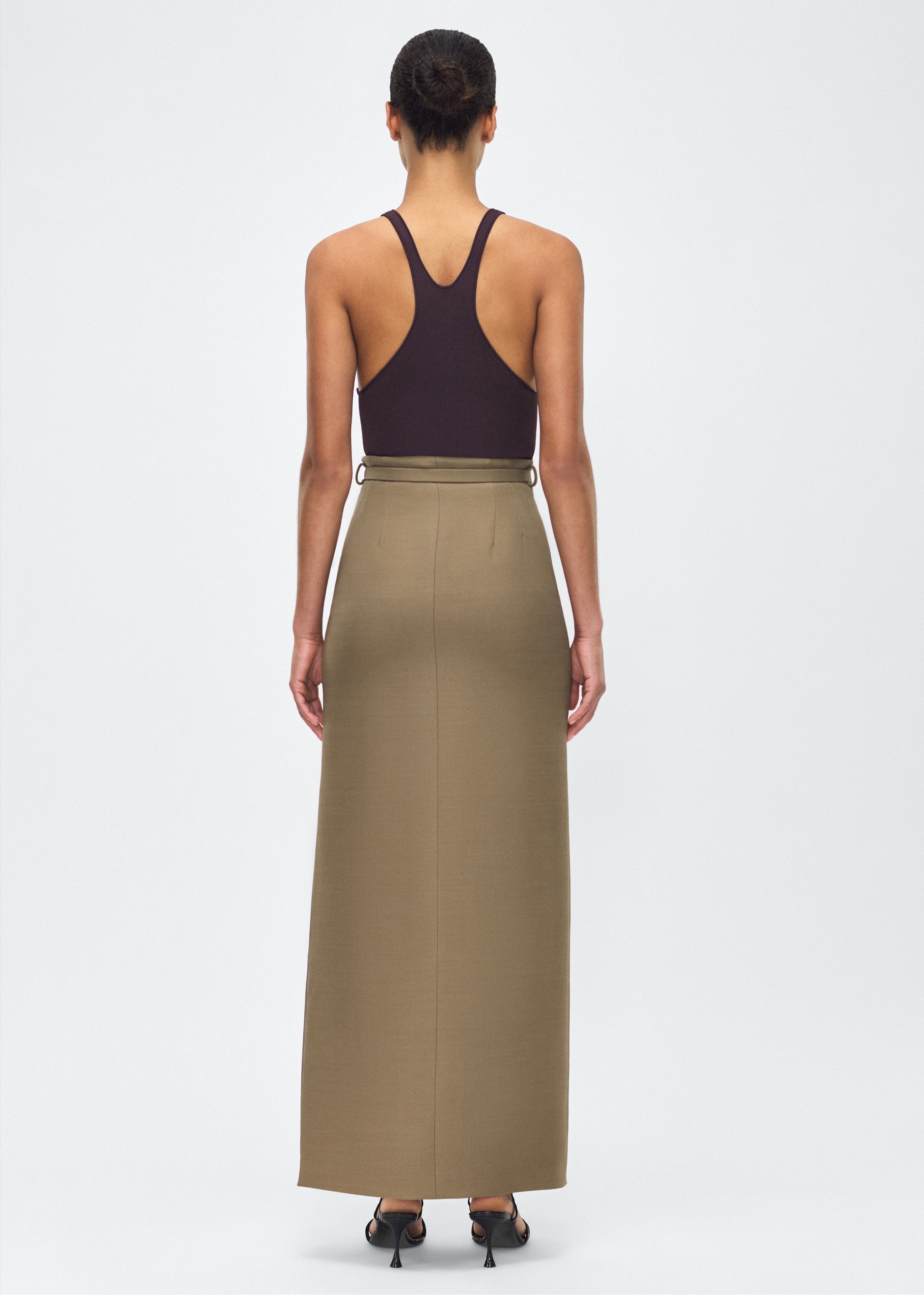Model facing back – Leto Skirt, P25412SW, Military Green – Adam Lippes, P25 collection:The Leto Skirt offers an elevated pencil silhouette, crafted from luxurious silk wool in a sophisticated military green hue. The sleek design is accentuated by a fabric-covered belt, cinching the waist for a tailored fit.