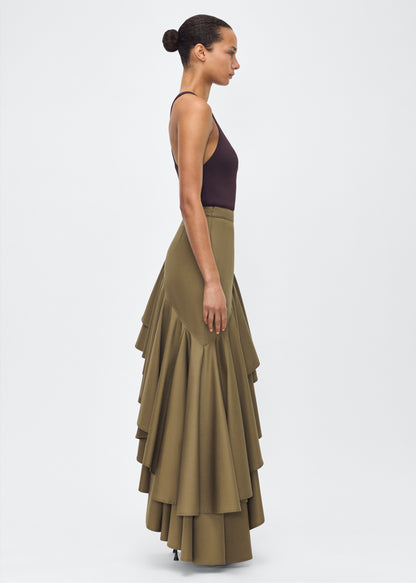 Model facing side – Cortes Skirt, P25413CT, Military Green – Adam Lippes, P25 collection: A new shape from the atelier, the Cortés Skirt is crafted from cotton twill in a rich military green hue. Featuring a dramatic asymmetrical ruffle, this skirt adds dynamic movement and texture. The high-waisted fit and expertly placed ruffles make it a statement piece that effortlessly transitions from day to night.