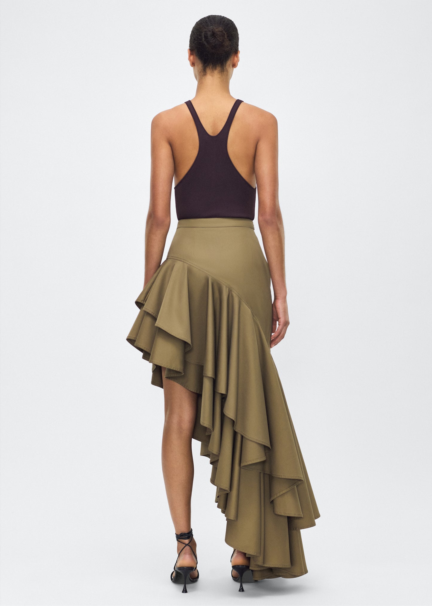 Model facing back – Cortes Skirt, P25413CT, Military Green – Adam Lippes, P25 collection: A new shape from the atelier, the Cortés Skirt is crafted from cotton twill in a rich military green hue. Featuring a dramatic asymmetrical ruffle, this skirt adds dynamic movement and texture. The high-waisted fit and expertly placed ruffles make it a statement piece that effortlessly transitions from day to night.