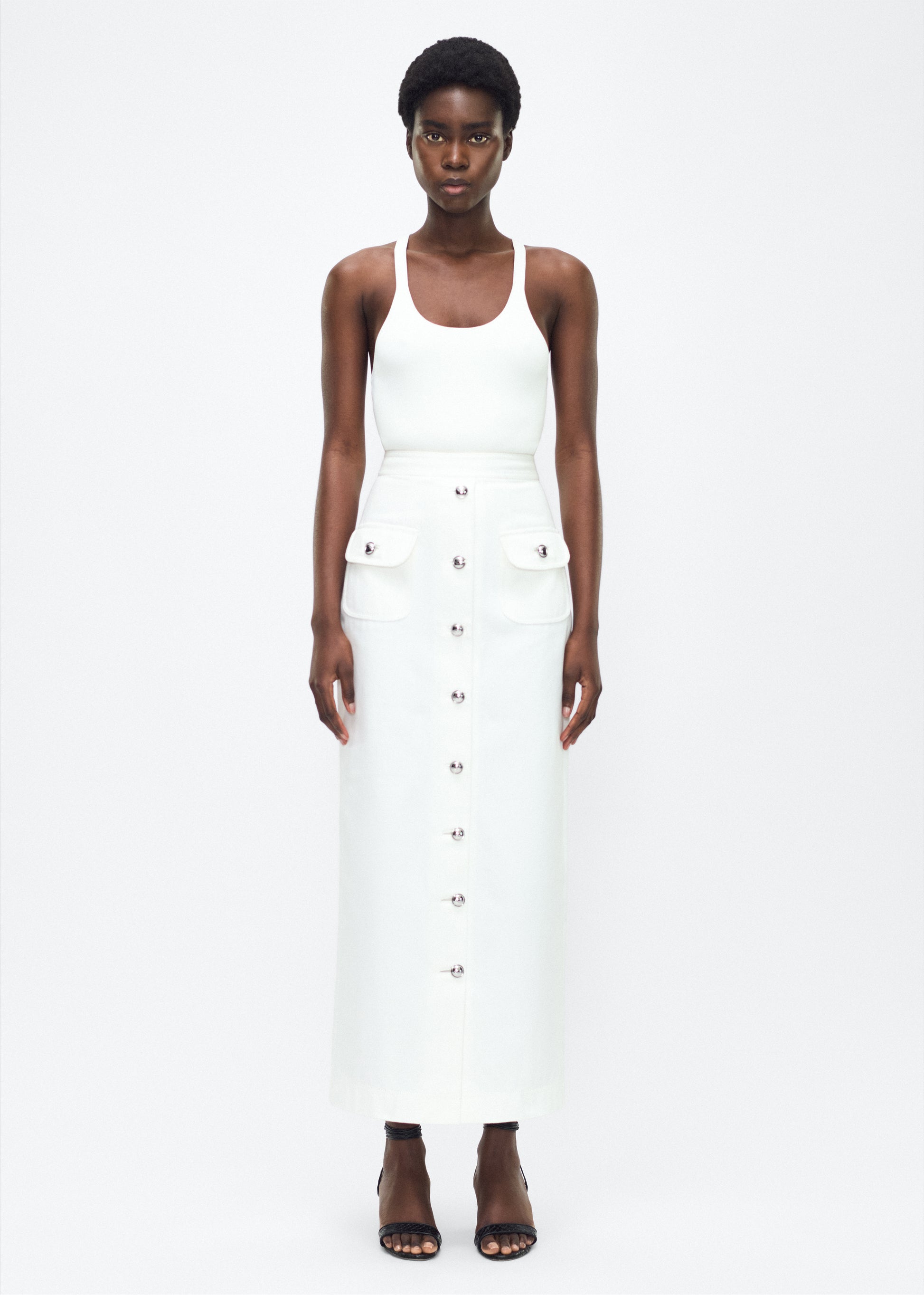 Image of model facing the front – Dakota Skirt, P25417WN, White – Adam Lippes, P25 collection: This classic skirt, crafted from Italian white denim, features elegant silver buttons cast with our iconic diamond monogram along the center front and functional front pockets. Featuring an invisible zipper for elegant wear, the tailored waist creates a defined silhouette. Pair with the Remo Top in White Denim for a complete look.