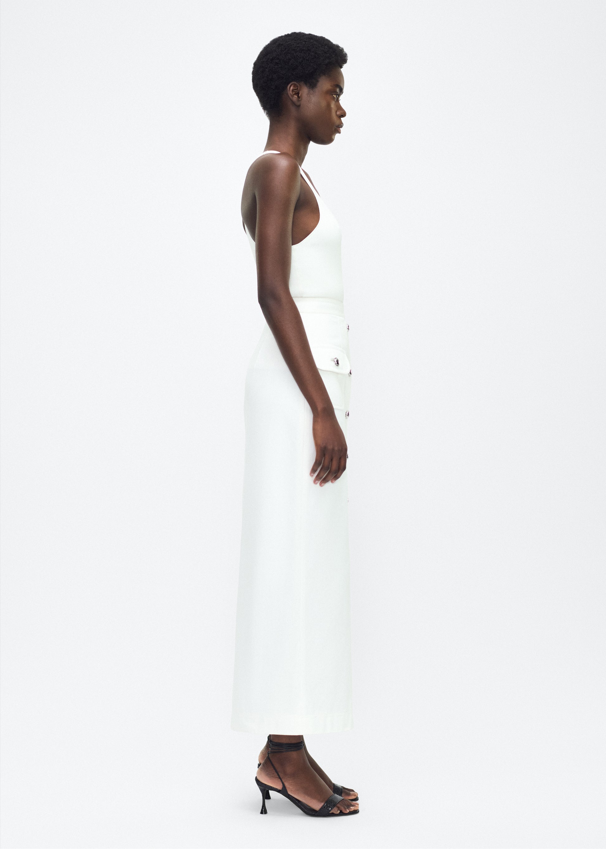 Image of model facing the side – Dakota Skirt, P25417WN, White – Adam Lippes, P25 collection: This classic skirt, crafted from Italian white denim, features elegant silver buttons cast with our iconic diamond monogram along the center front and functional front pockets. Featuring an invisible zipper for elegant wear, the tailored waist creates a defined silhouette. Pair with the Remo Top in White Denim for a complete look.