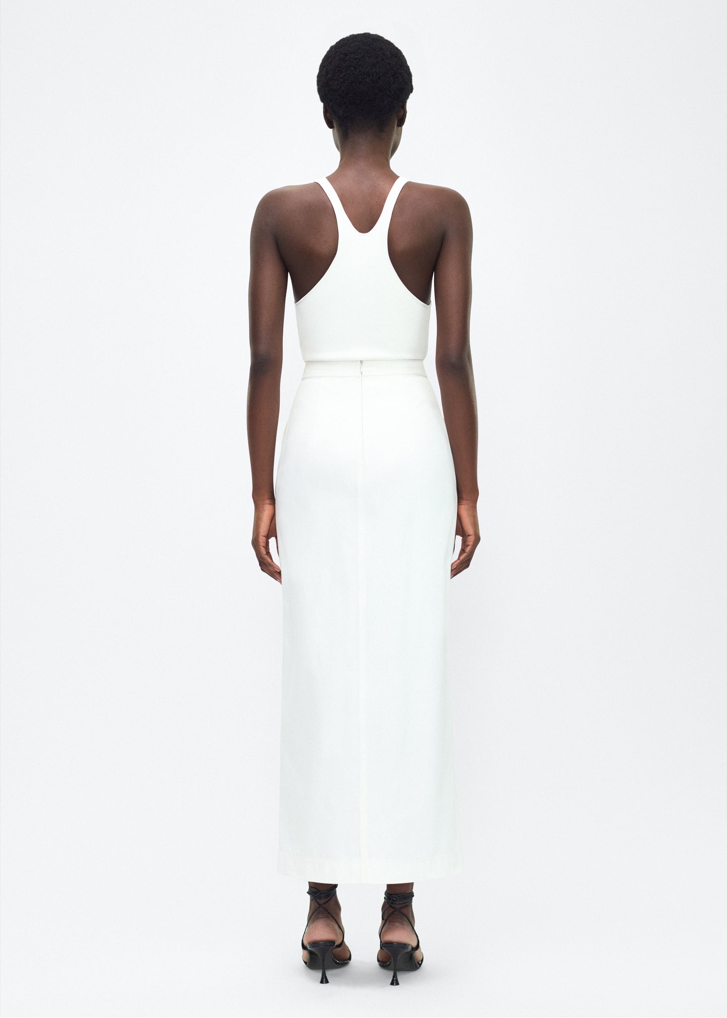 Image of model facing the back – Dakota Skirt, P25417WN, White – Adam Lippes, P25 collection: This classic skirt, crafted from Italian white denim, features elegant silver buttons cast with our iconic diamond monogram along the center front and functional front pockets. Featuring an invisible zipper for elegant wear, the tailored waist creates a defined silhouette. Pair with the Remo Top in White Denim for a complete look.