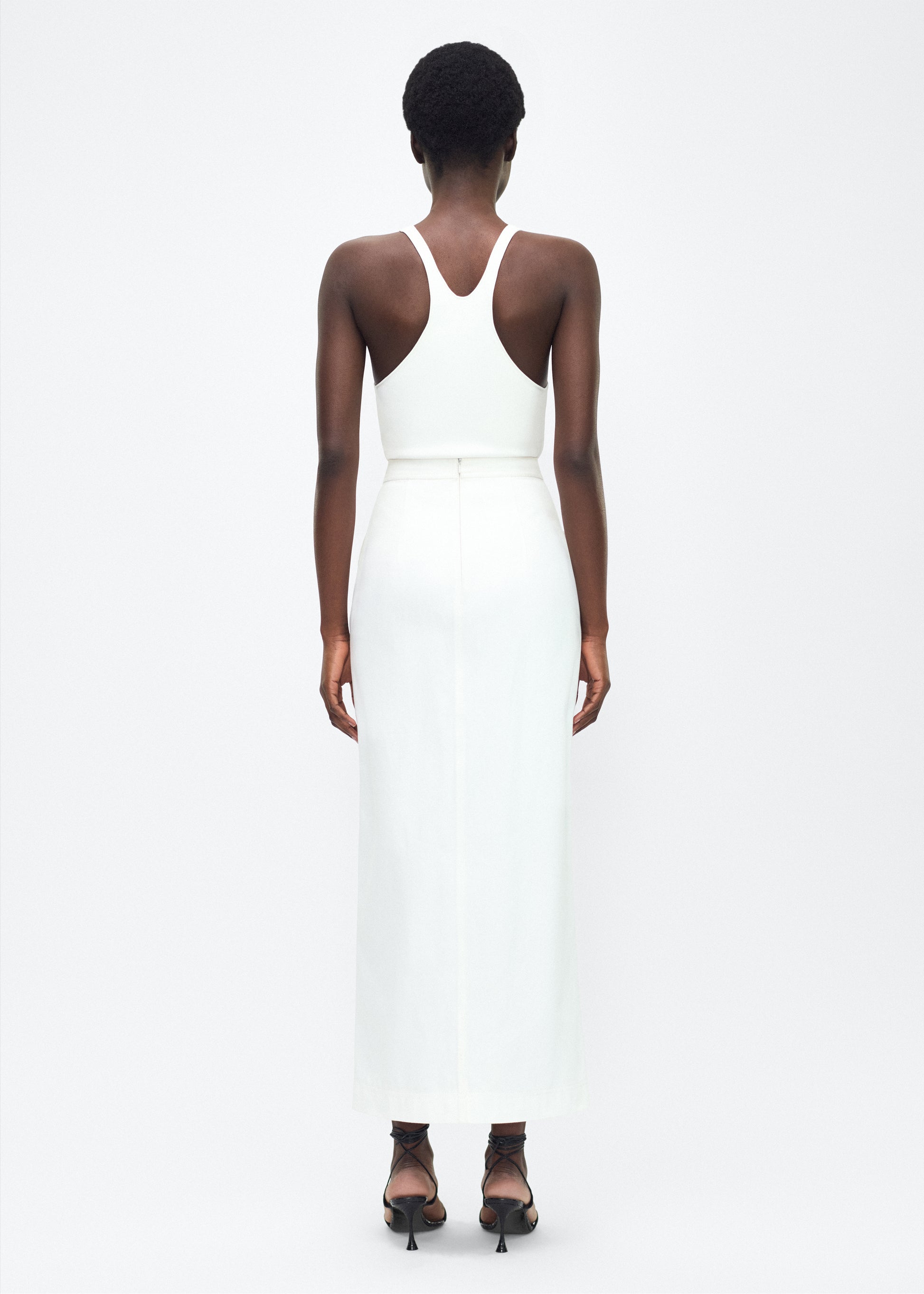 Image of model facing the back – Dakota Skirt, P25417WN, White – Adam Lippes, P25 collection: This classic skirt, crafted from Italian white denim, features elegant silver buttons cast with our iconic diamond monogram along the center front and functional front pockets. Featuring an invisible zipper for elegant wear, the tailored waist creates a defined silhouette. Pair with the Remo Top in White Denim for a complete look.