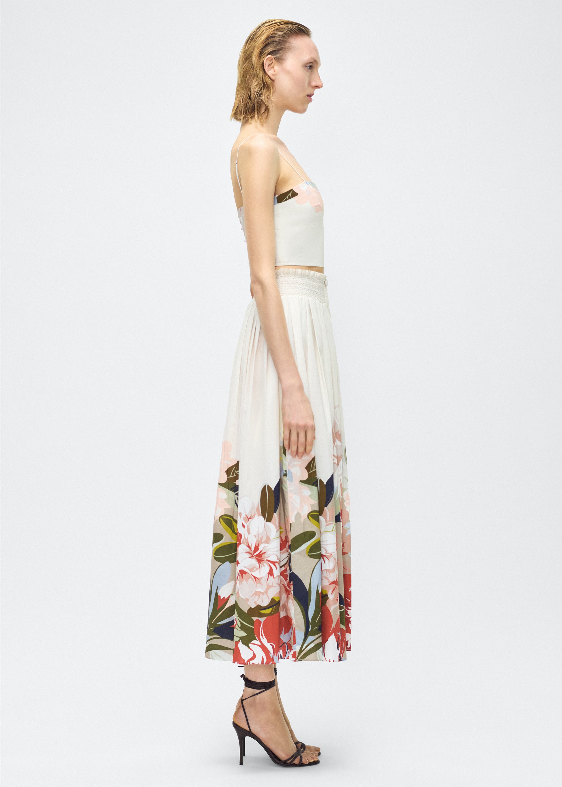 Image of model side- Lovisa Skirt, P25418PV, Champagne Multi – Adam Lippes, P25 collection: A signature shape from the atelier, the Lovisa Skirt is crafted from airy printed voile featuring a vibrant Sicily-inspired floral motif. Designed with a smocked waistband for a comfortable fit, the skirt is finished with fabric-covered buttons down the center front. Pair with the Bex Top in Printed Voile for a coordinated look.